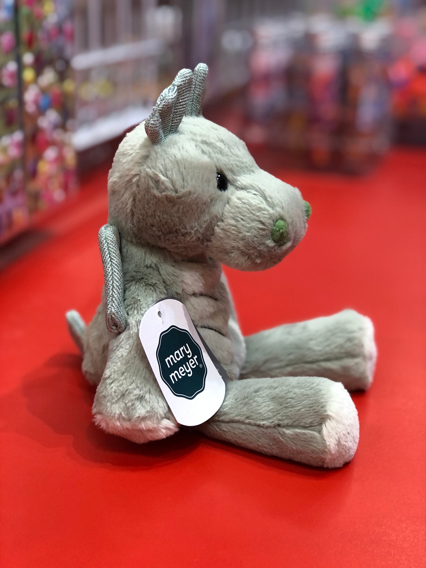 Soft plush Mary Meyer Marshmallow Zoo dragon toy with silver ears and wings, sitting on red surface, tag visible