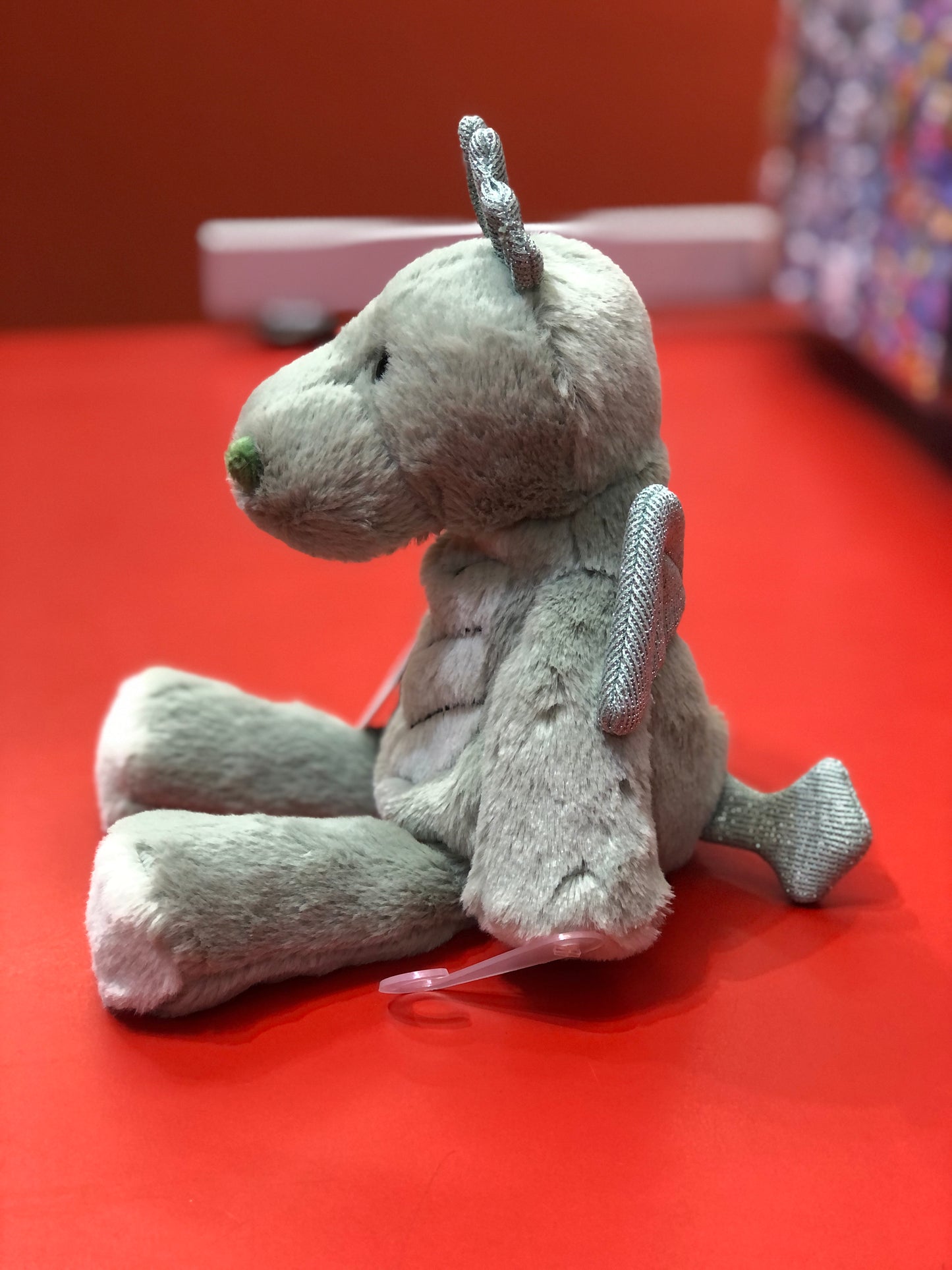 Soft plush Marshmallow Zoo dragon toy sitting on red surface, with silver accents and green nose, ready for cuddling