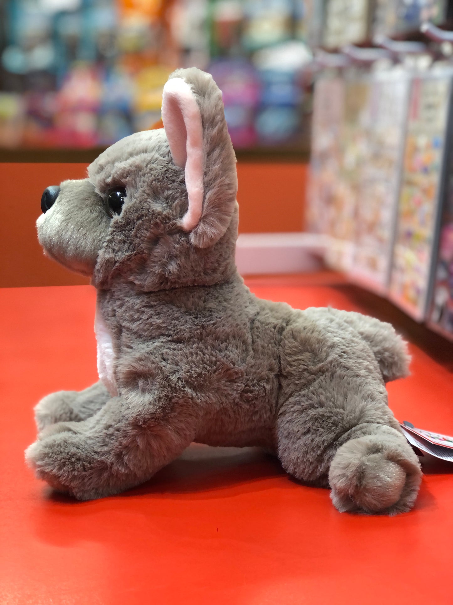 Lilac French Bulldog plush toy with oversized ears and bright eyes, sitting on red surface in toy store display