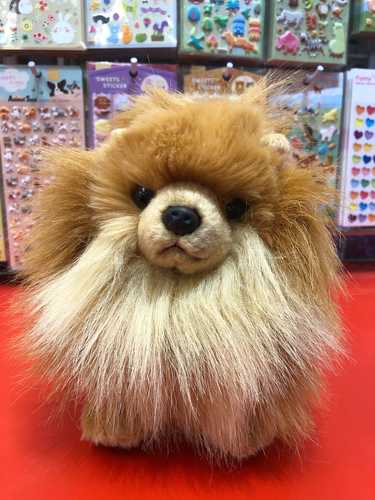 Fluffy Pomeranian plush toy with tan and light brown fur, pointed ears, on red display with colorful stickers in background