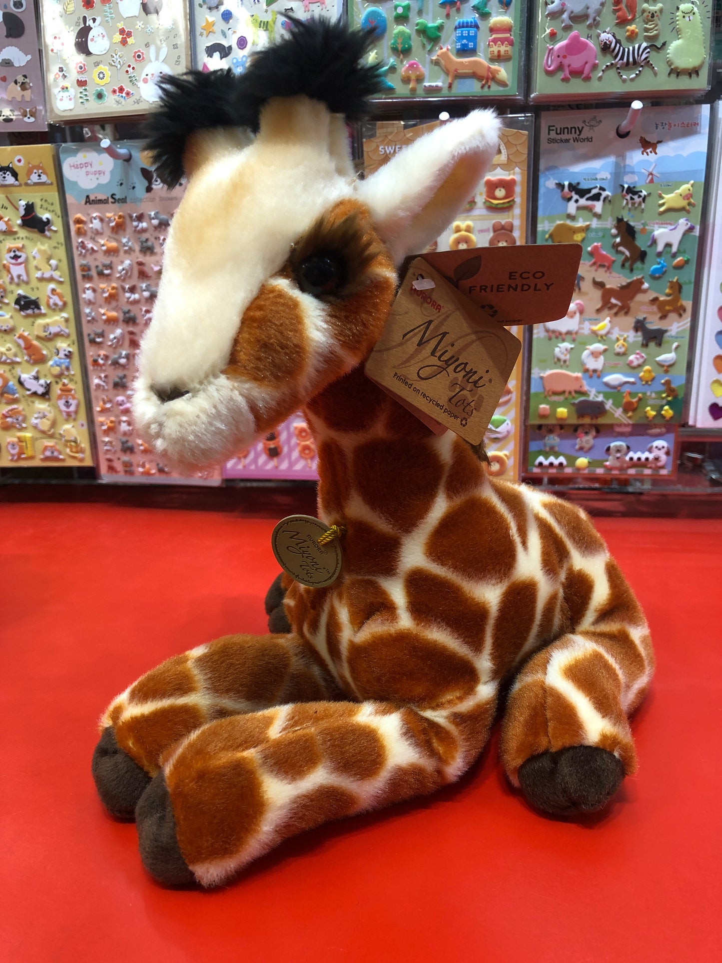 Cuddly Aurora Miyoni Tots Giraffe Calf plush toy with realistic markings, displayed on red surface against colorful animal sticker background