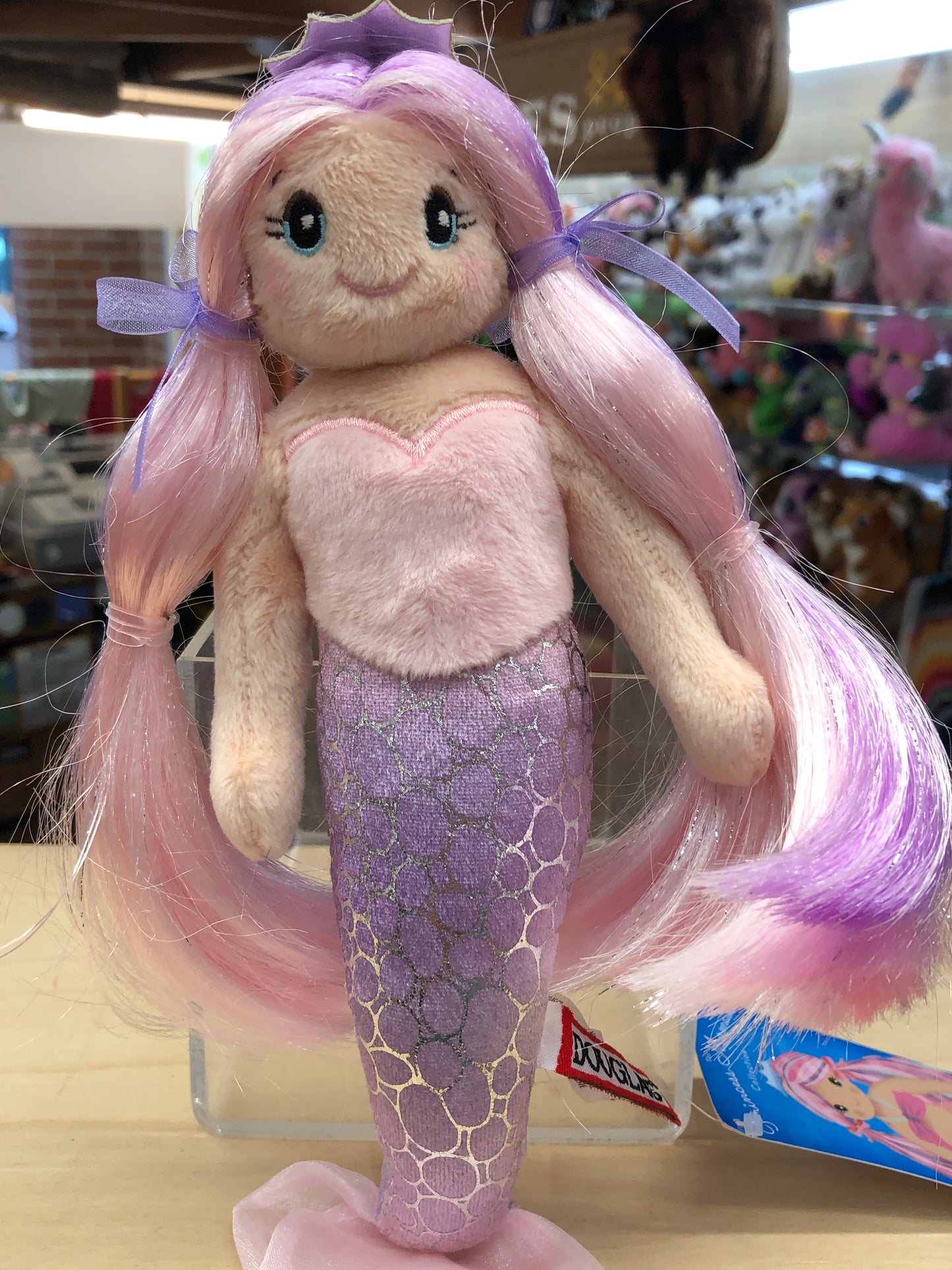 Douglas Princess Mermaid plush toy with pink hair, purple crown, and sparkly tail, displayed in store