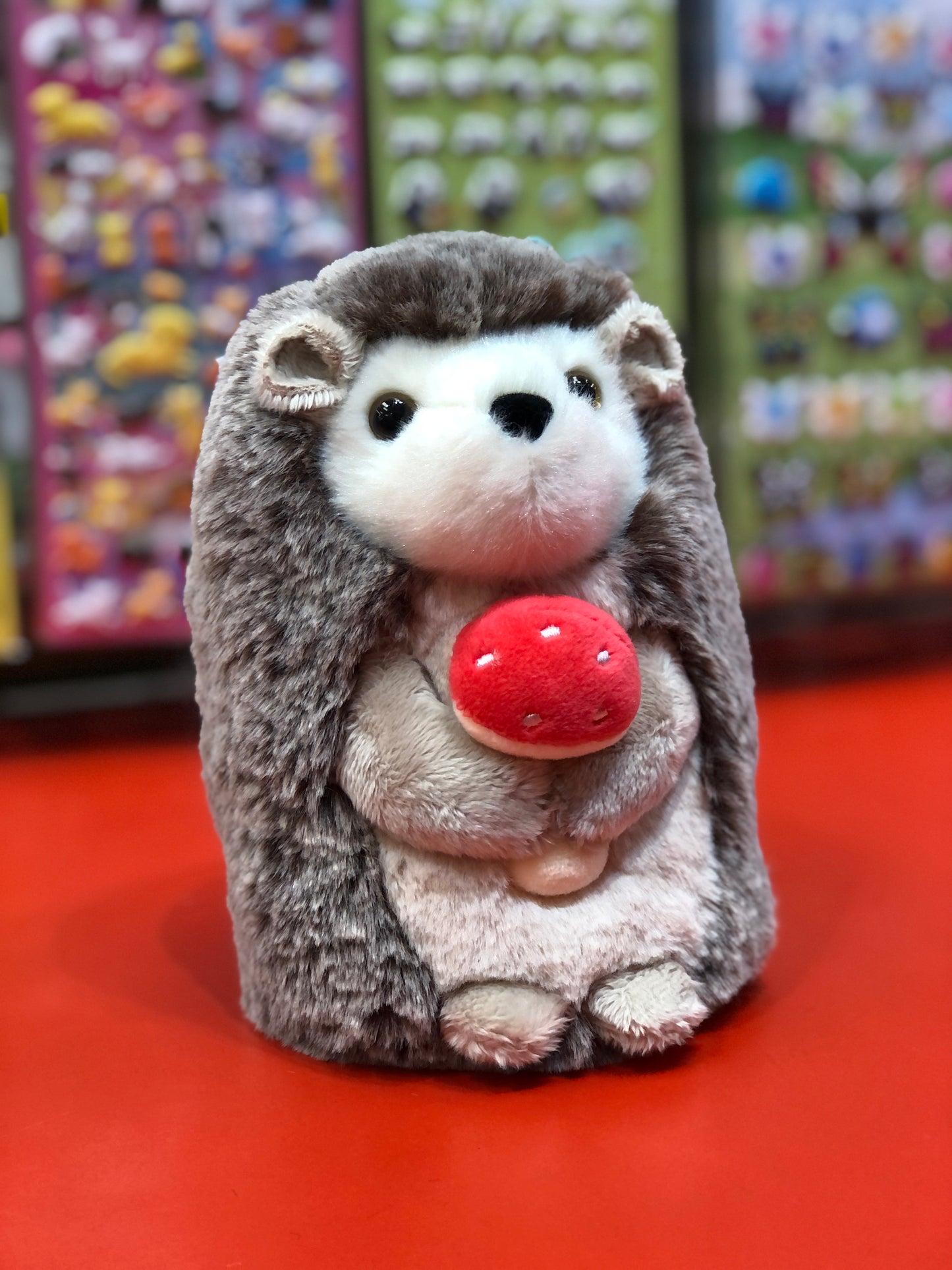 Adorable Douglas Stuey hedgehog plush toy holding red mushroom, displayed on red surface with colorful background