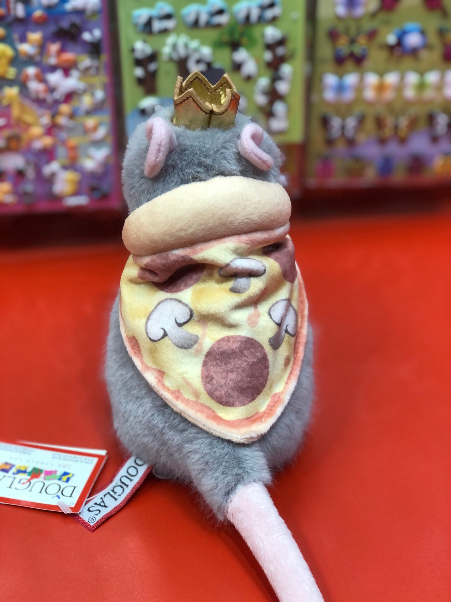 Cute plush rat toy wearing golden crown and pizza-slice cape, displayed on red surface with colorful background