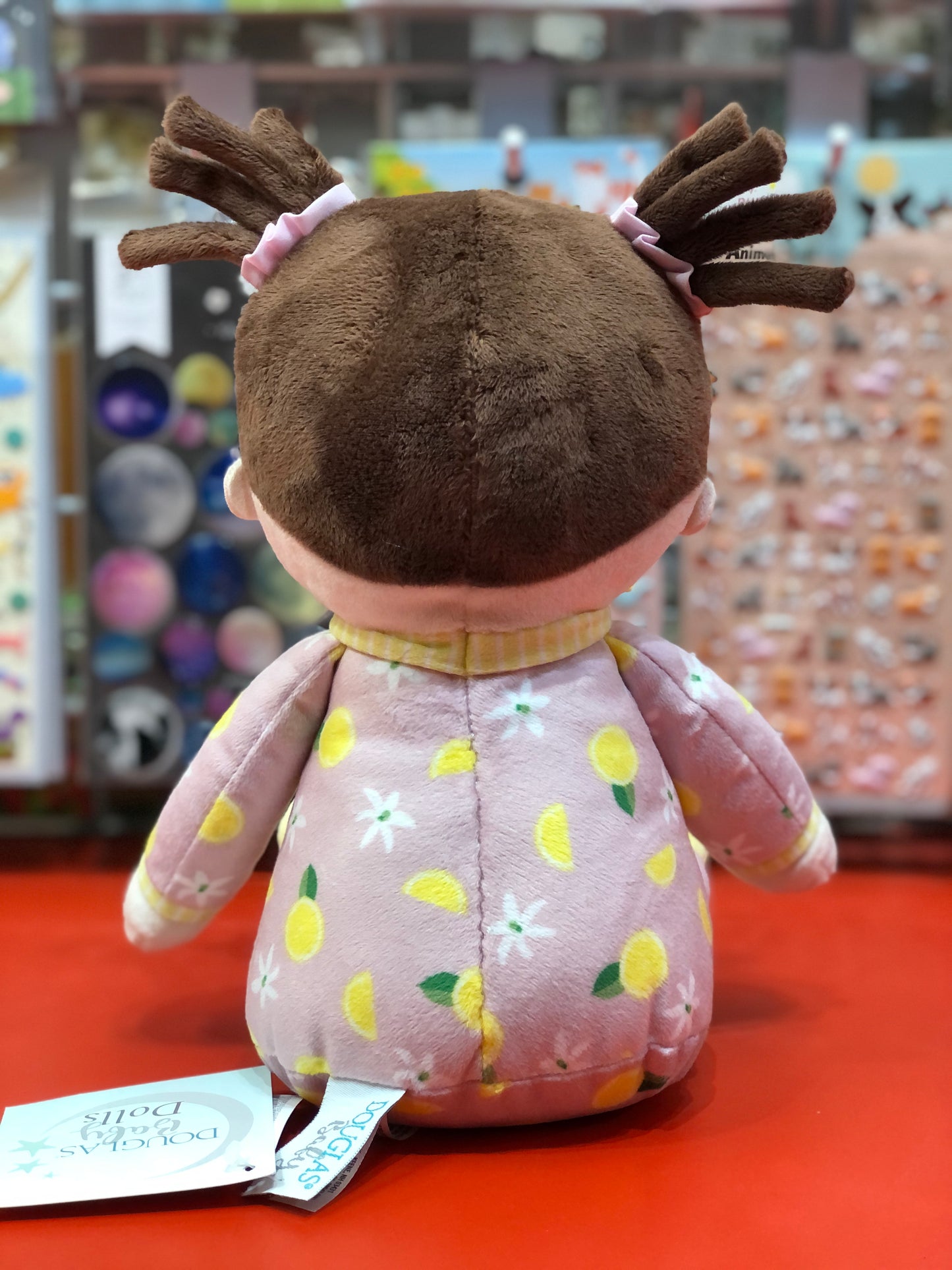 Lainey Lemon Doll plush with pink lemon-patterned PJs, pigtails, and gingham trim, shown from back on red display shelf