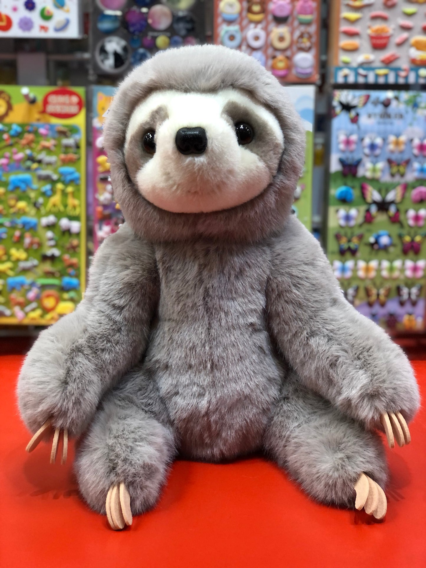 Cuddly Douglas Super Lizzie Soft Sloth plush toy with realistic features, sitting on red surface in colorful toy store display