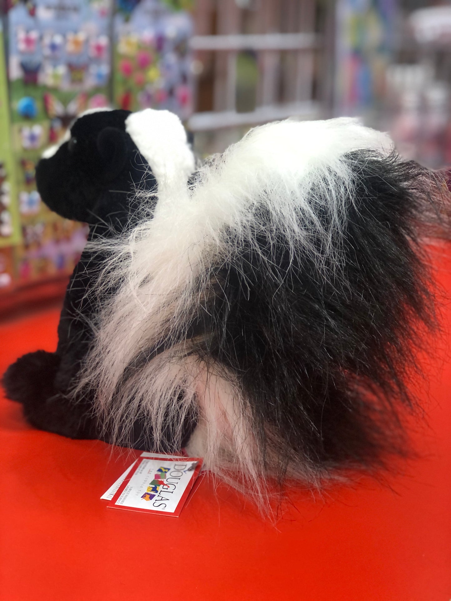 Douglas Odie Skunk plush toy with black and white fur, fluffy tail, seated on red surface, colorful background