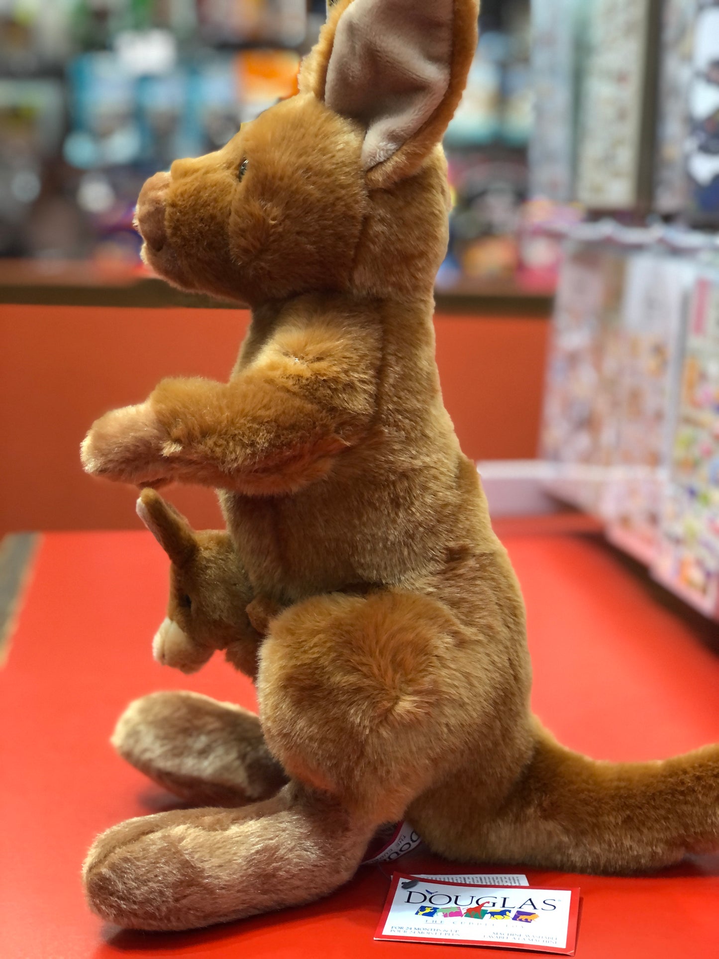 Plush kangaroo toy with joey puppet in pouch, sitting on red surface, Douglas brand tag visible