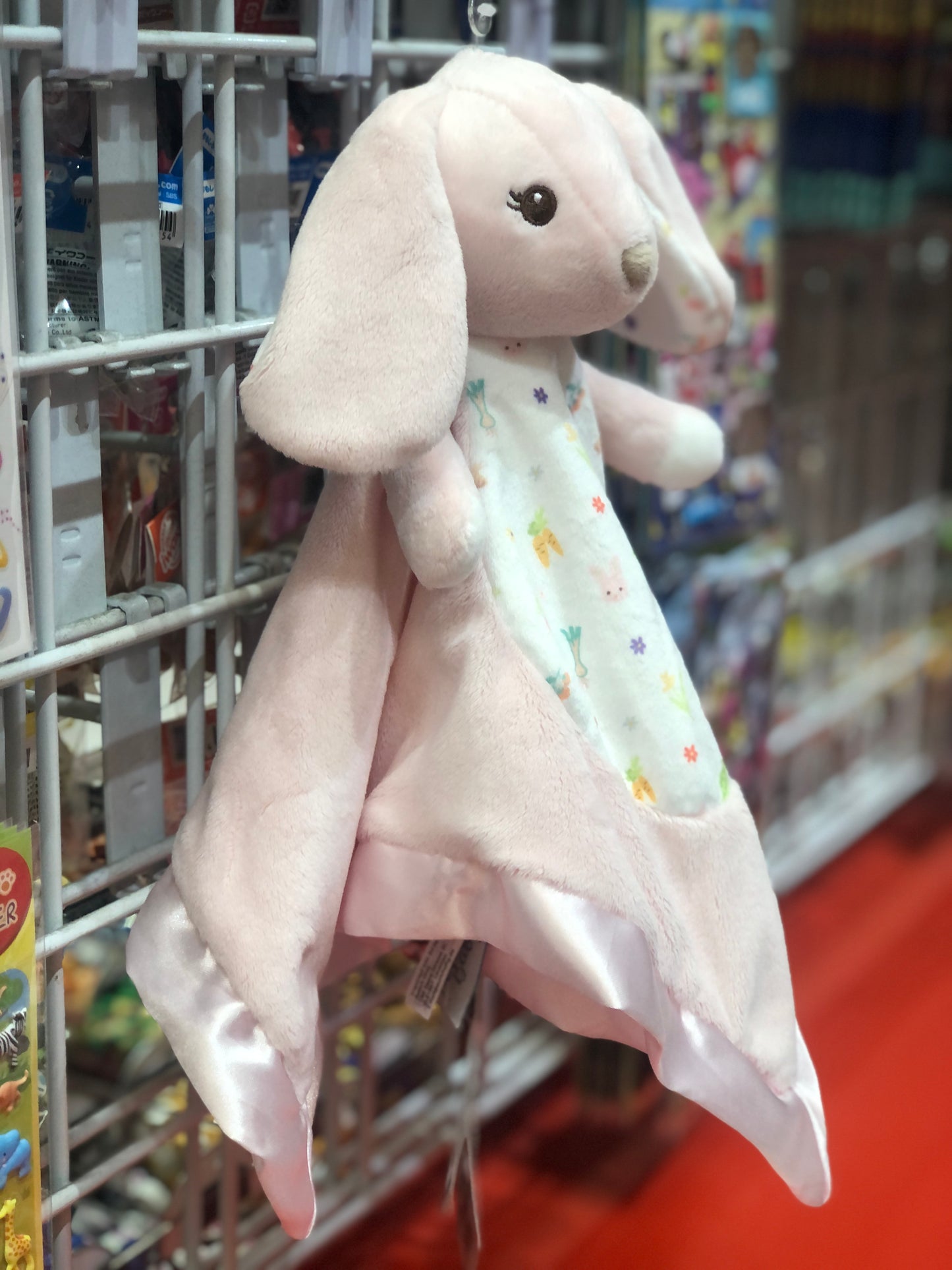 Pink bunny plush toy with satin trim and floral print blanket, displayed on store shelf for baby cuddling and comfort