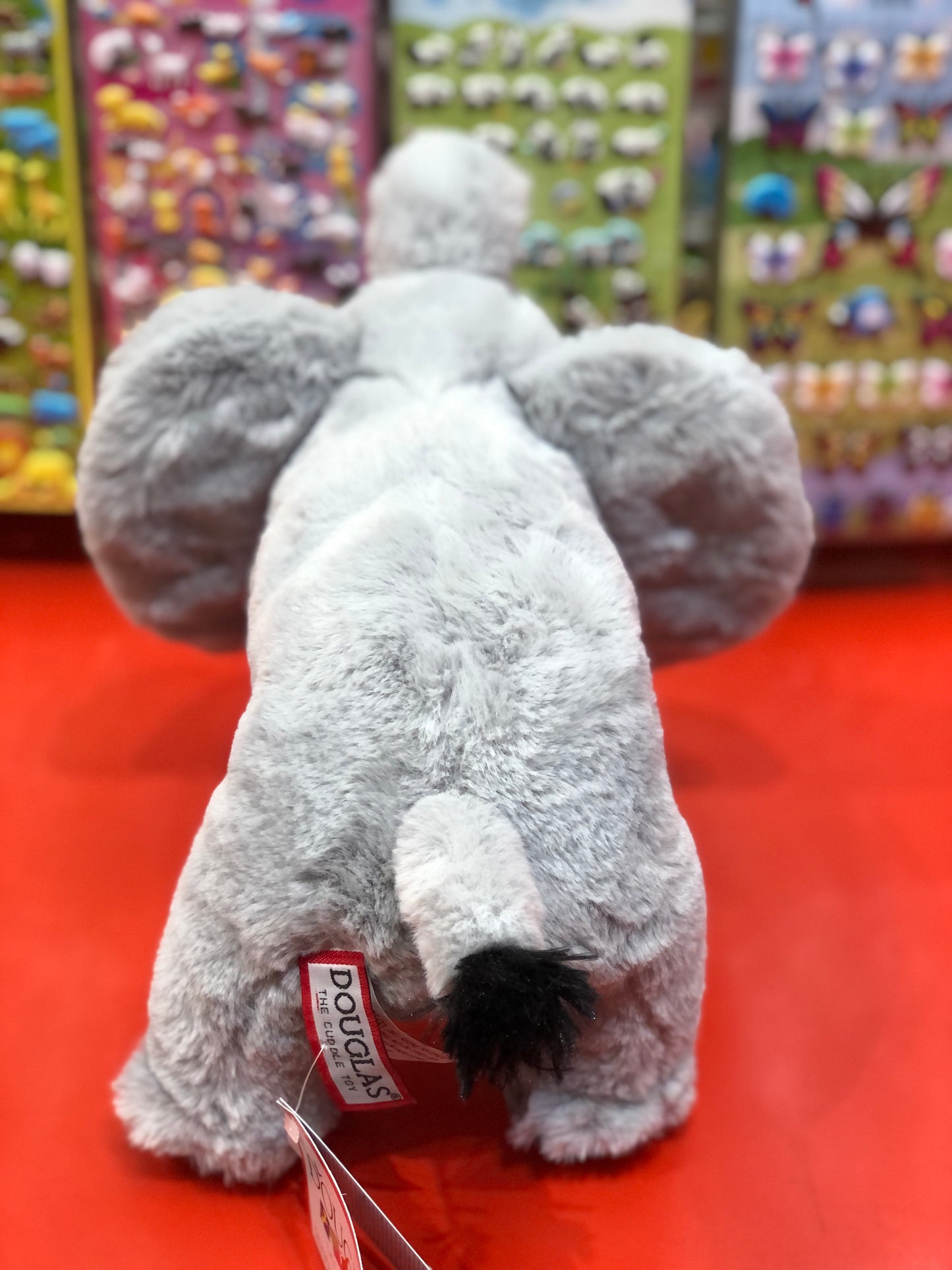 Soft gray Douglas Everlie elephant plush toy on red display, colorful stuffed animal display in background