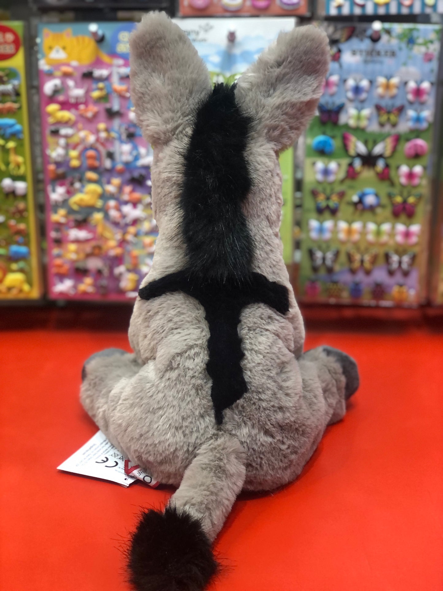 Soft gray and black plush donkey toy sitting on red surface with colorful toy displays in background