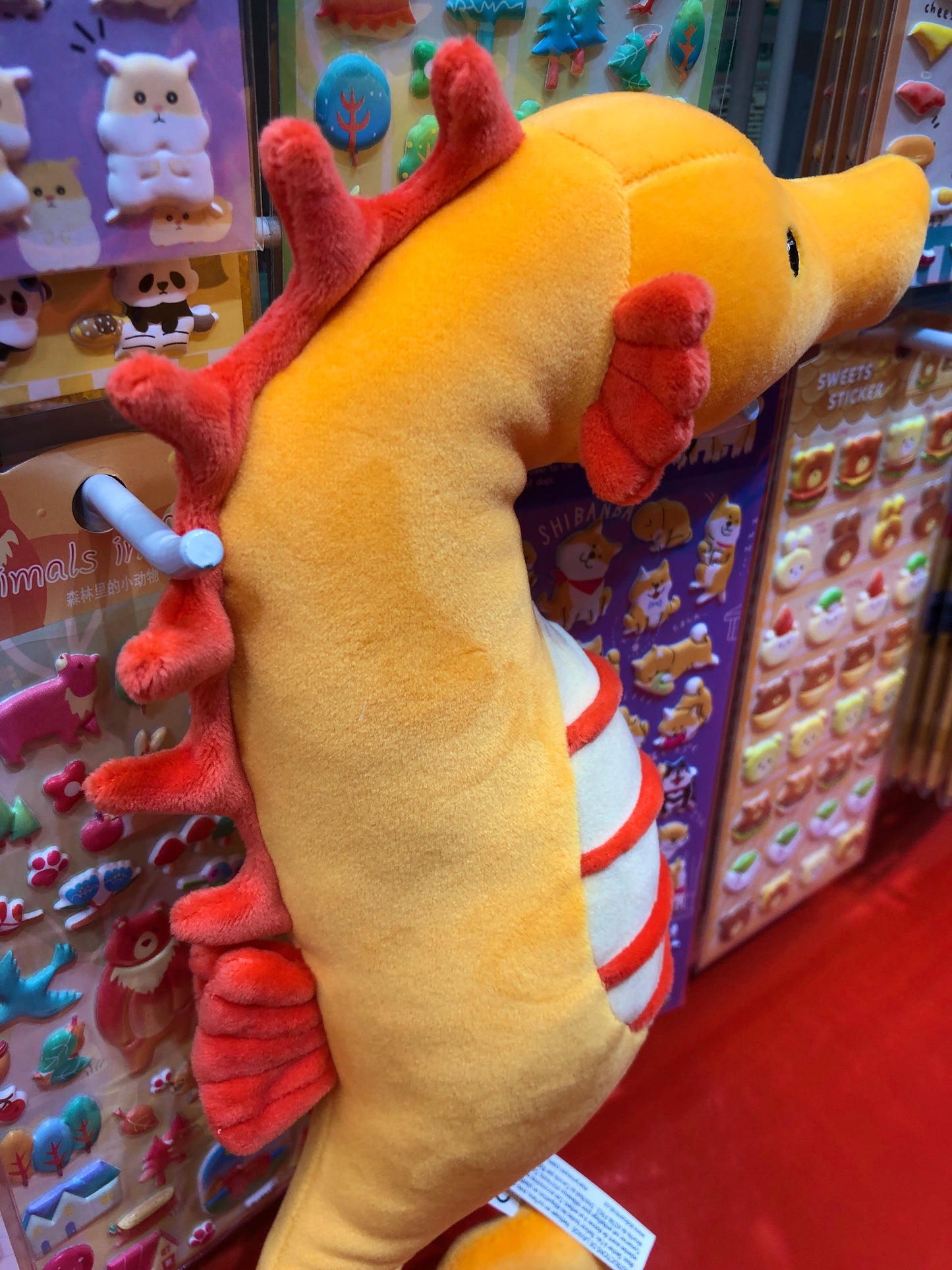 Soft yellow and red plush seahorse toy with embroidered eyes, surrounded by colorful animal stickers and toys in store display