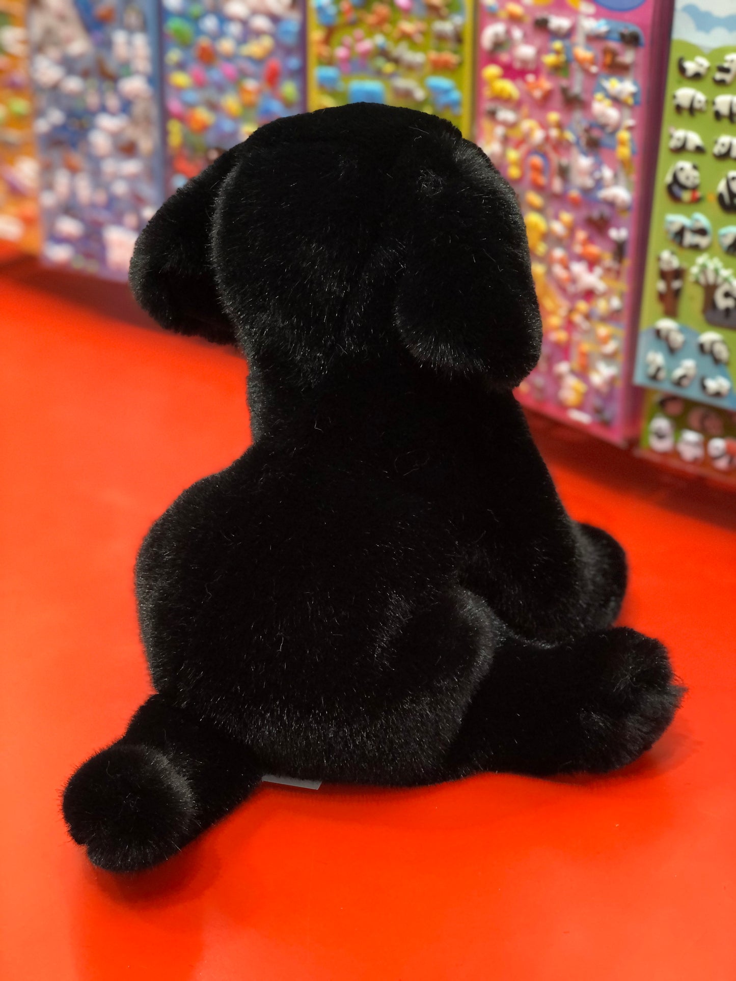 Soft black Labrador plush toy sitting on red surface, colorful toy display in background