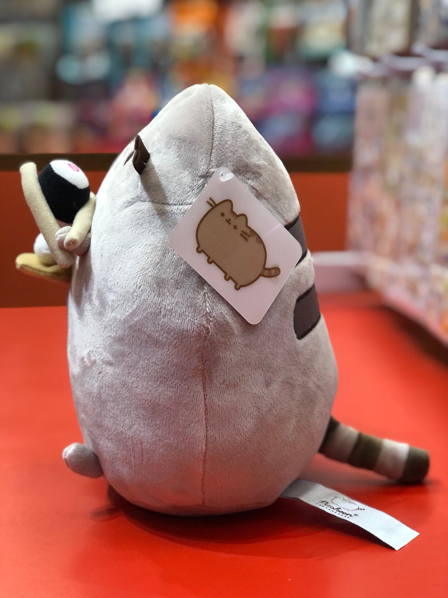 Pusheen Snackable Sushi plush toy holding chopsticks, displayed on red surface with product tag visible