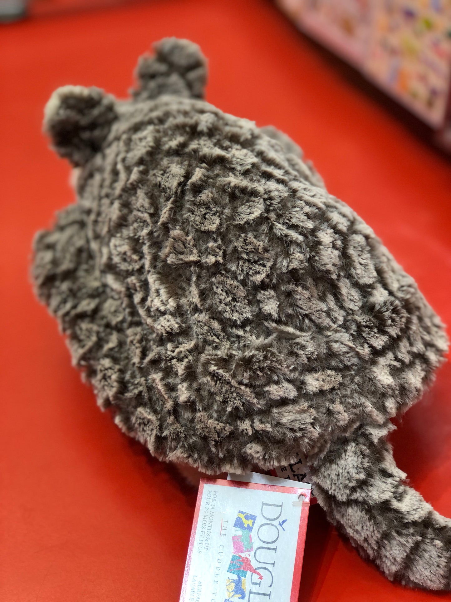Soft gray Douglas Austin armadillo plush toy with textured fur on red background, showcasing whimsical design and tag