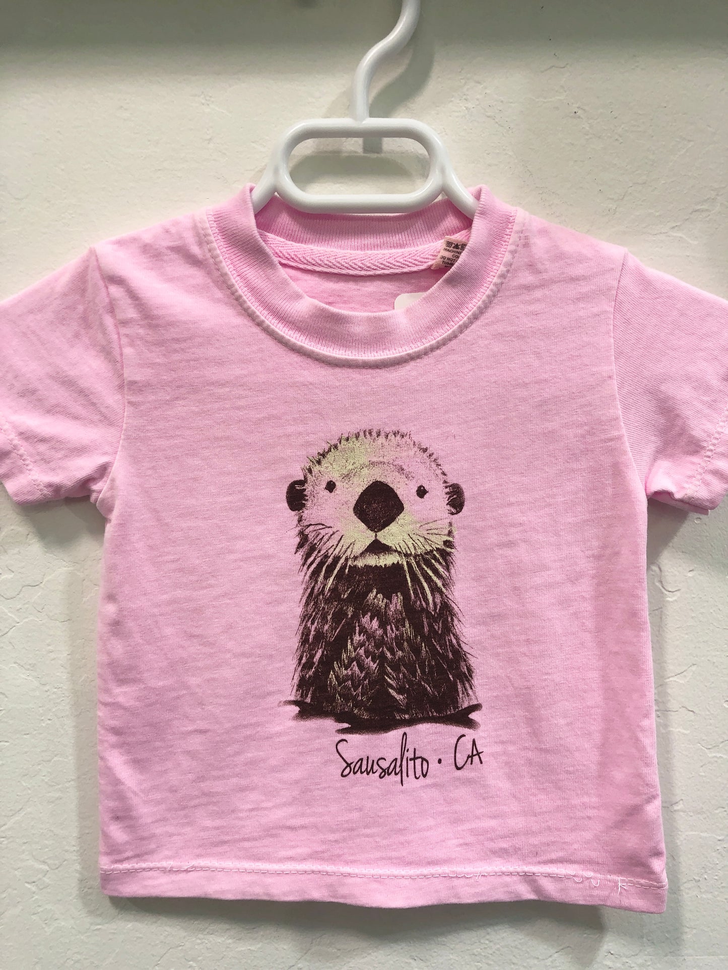 Pink infant t-shirt with cute sea otter illustration and "Sausalito CA" text, hanging on white hanger