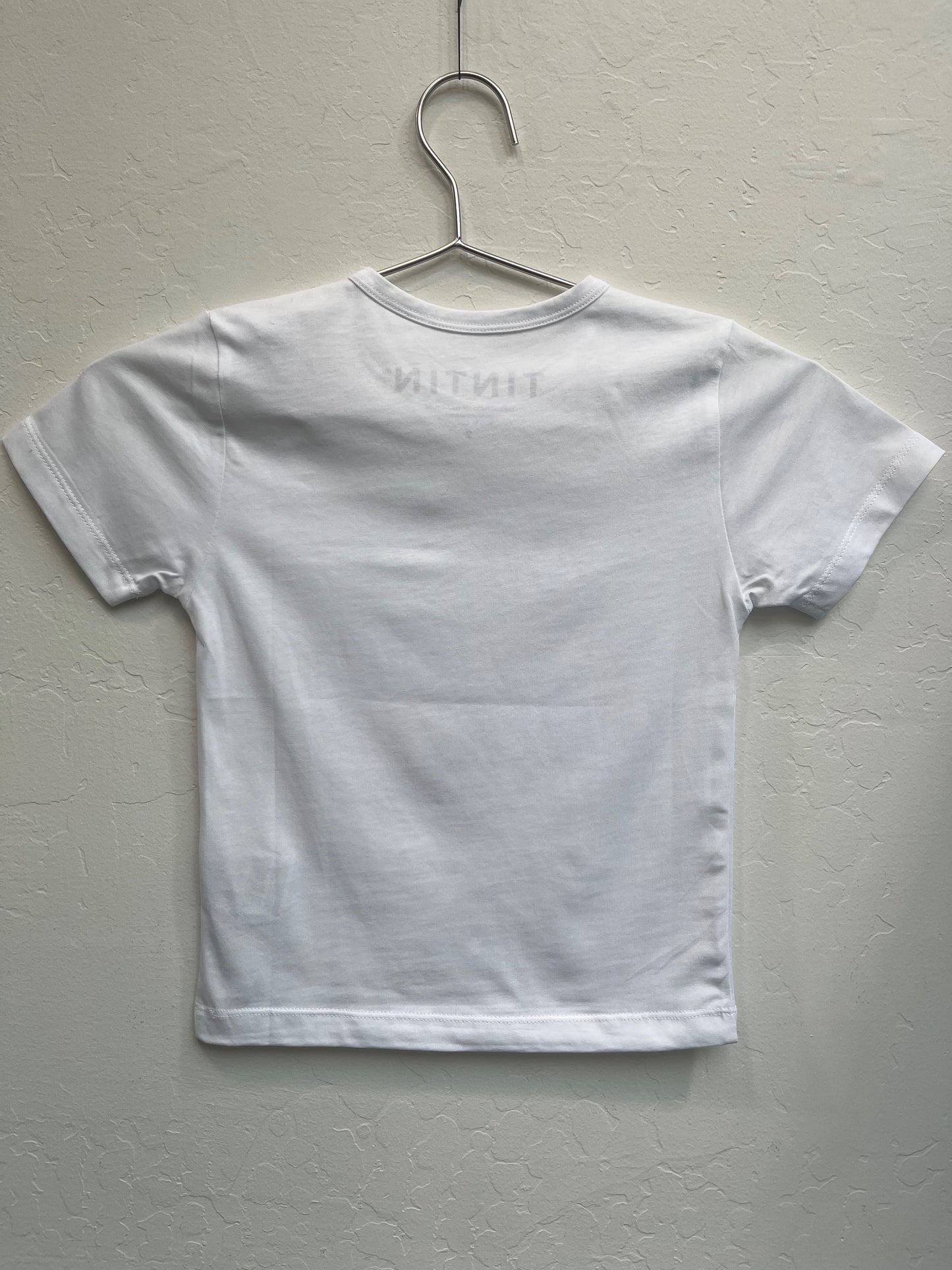 White kids' Tintin t-shirt with scoop neck hanging on metal hanger, showing back view with faint "TINTIN" text visible