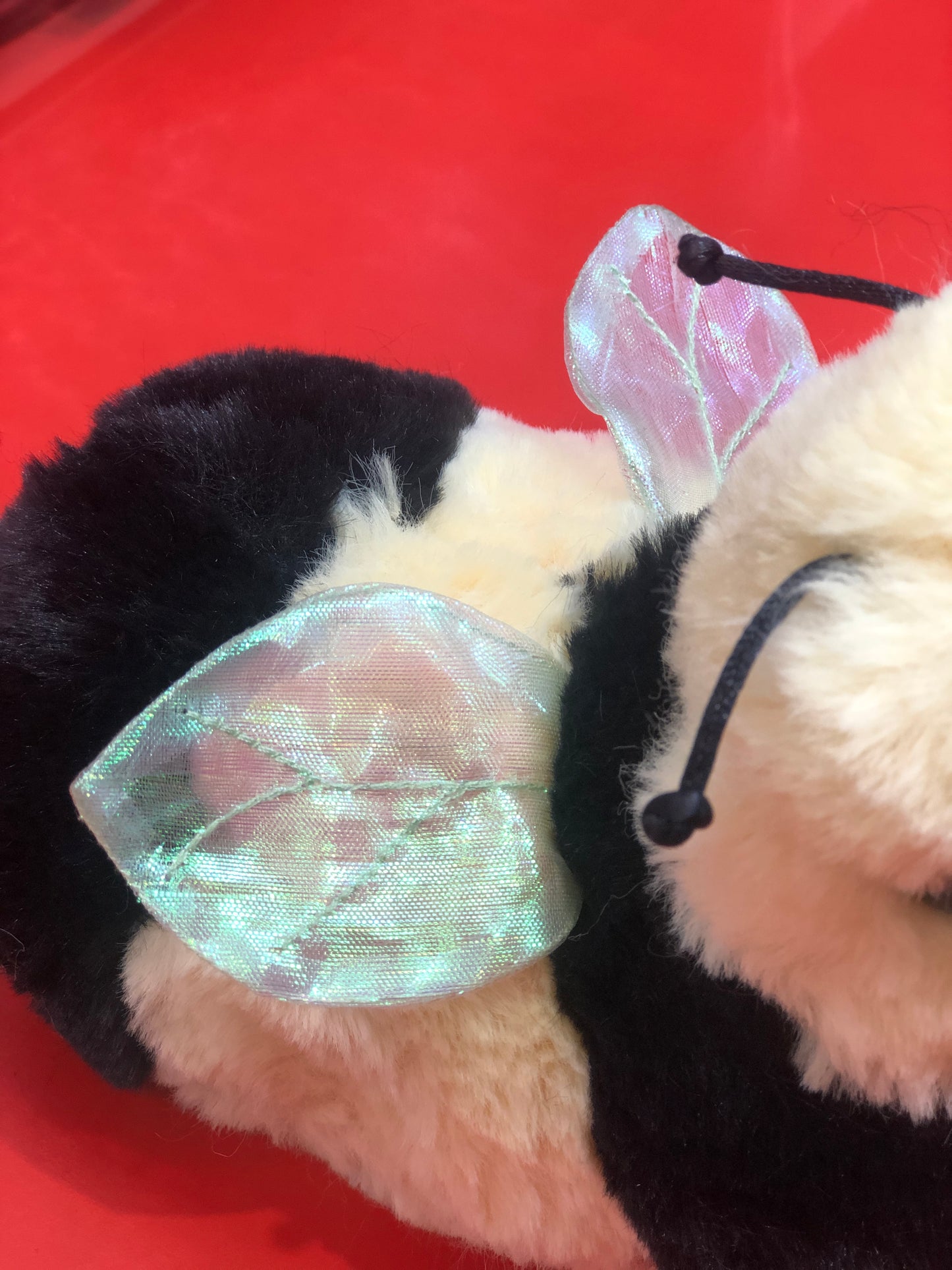 Soft bee plush toy with black and cream fur, featuring iridescent wings against a red background