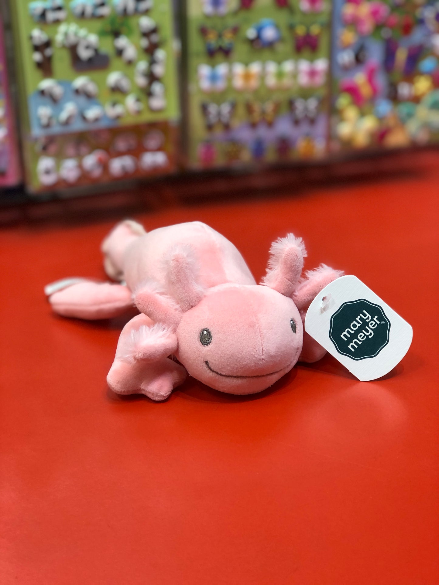 Soft pink Mary Meyer Smootheez axolotl plush toy with smiling face on red surface, product tag visible