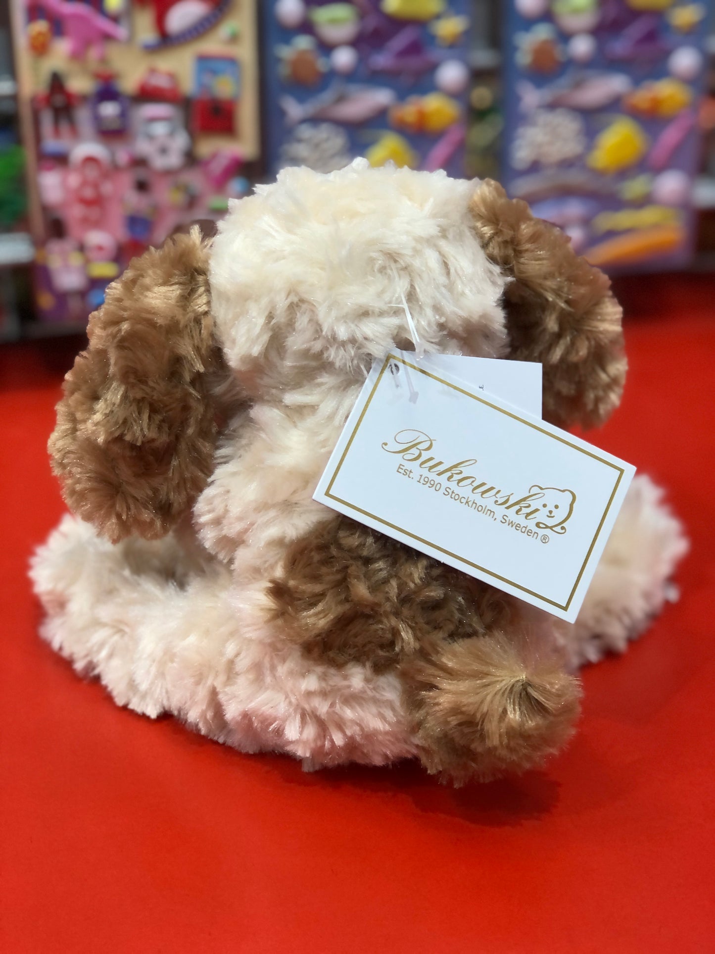 Cuddly Buddylina puppy plush with floppy ears and white and tan curly fur, sitting on red surface with Bukowski tag