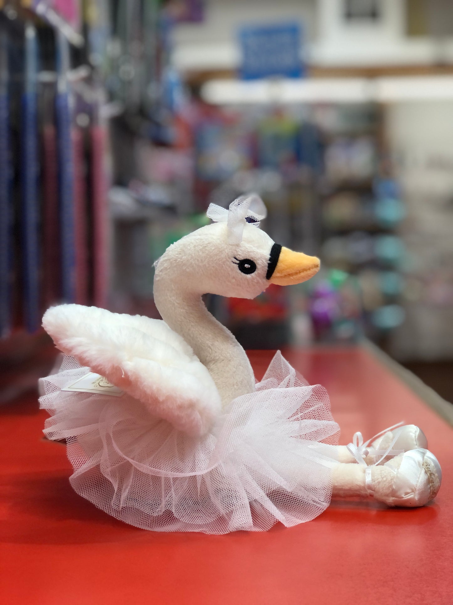 White swan ballerina plush toy with tulle skirt and ballet shoes, displayed on red surface in toy store