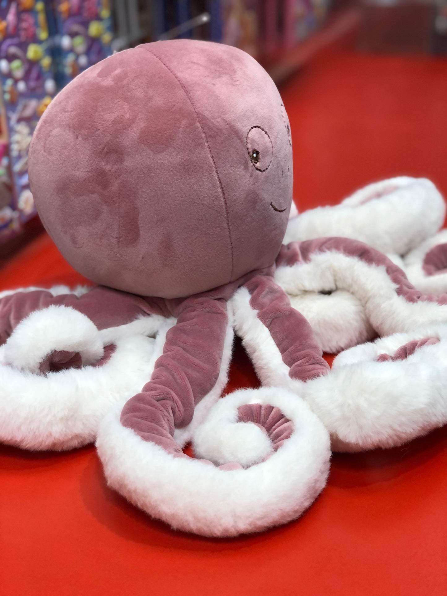 Cute pink octopus plush toy with soft white tentacles and smiling face, sitting on red surface in store display