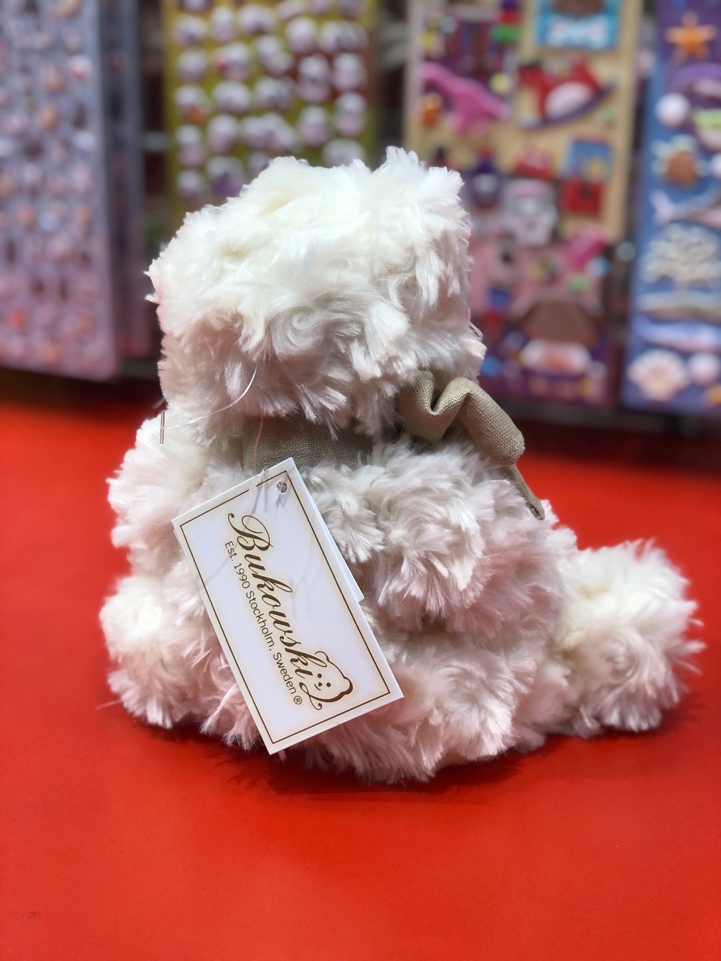 Cute cream-colored Bukowski Little Noah bear plush with tan bow, sitting on red surface, tag attached