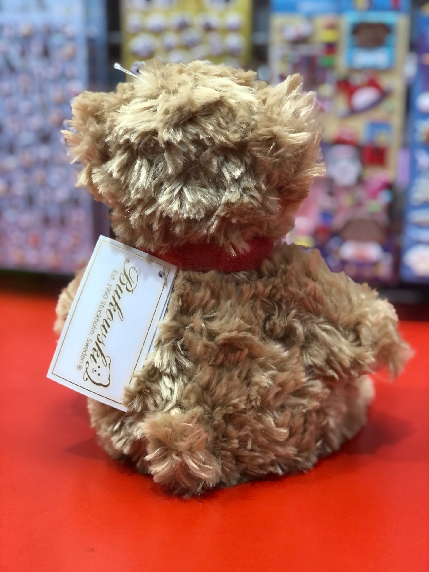 Bukowski Bears Clever Anton plush toy with curly fur and orange scarf, sitting on red surface with tag attached