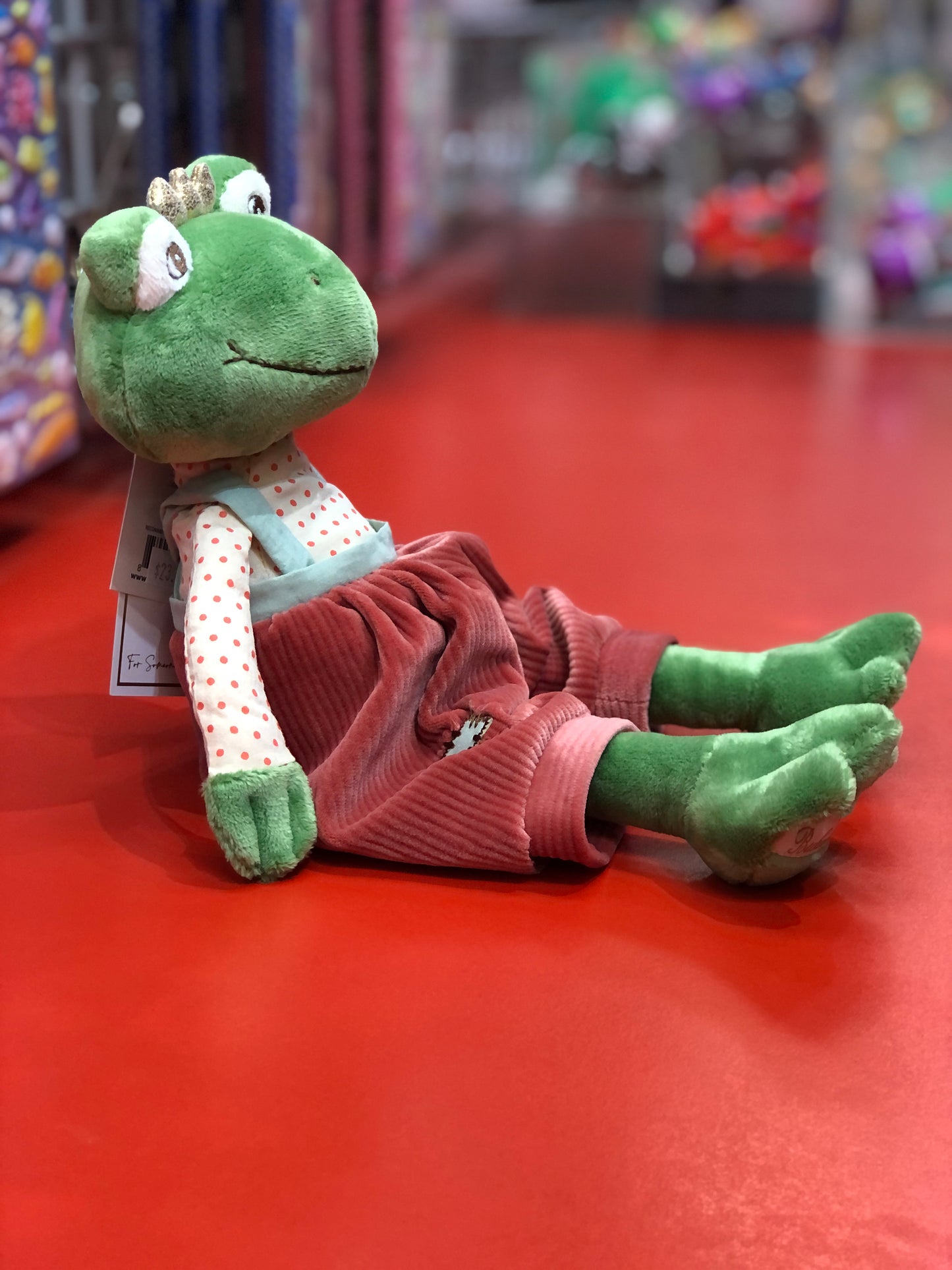 Bukowski Bears Prince Frog plush toy wearing polka-dot shirt and red overalls, sitting on red surface in toy store