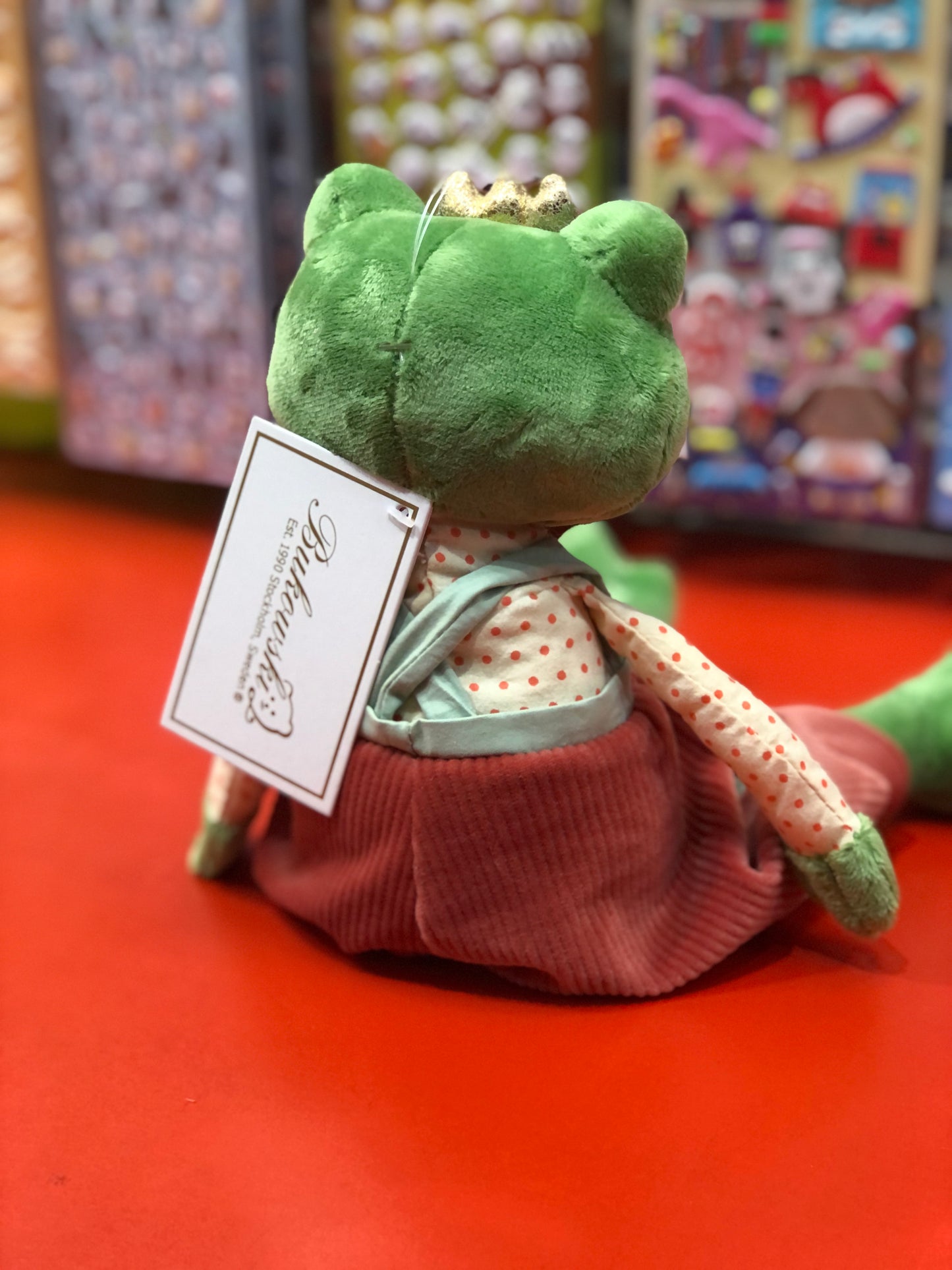 Bukowski Bears Prince Frog plush toy with golden crown, polka-dot shirt, and red overalls, displayed on red surface in toy store