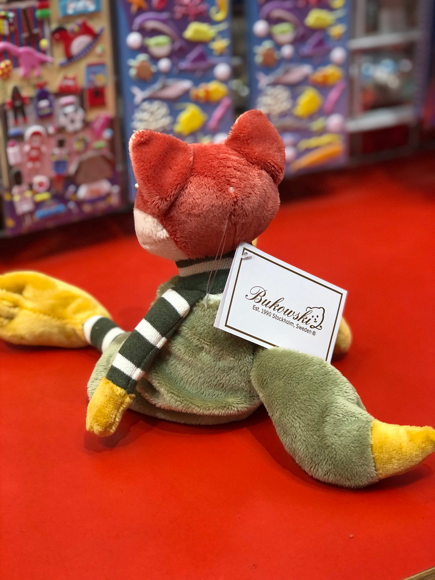 Bukowski Bears Foxy Fox plush toy with orange head, green body, and striped scarf, sitting on red surface in toy store