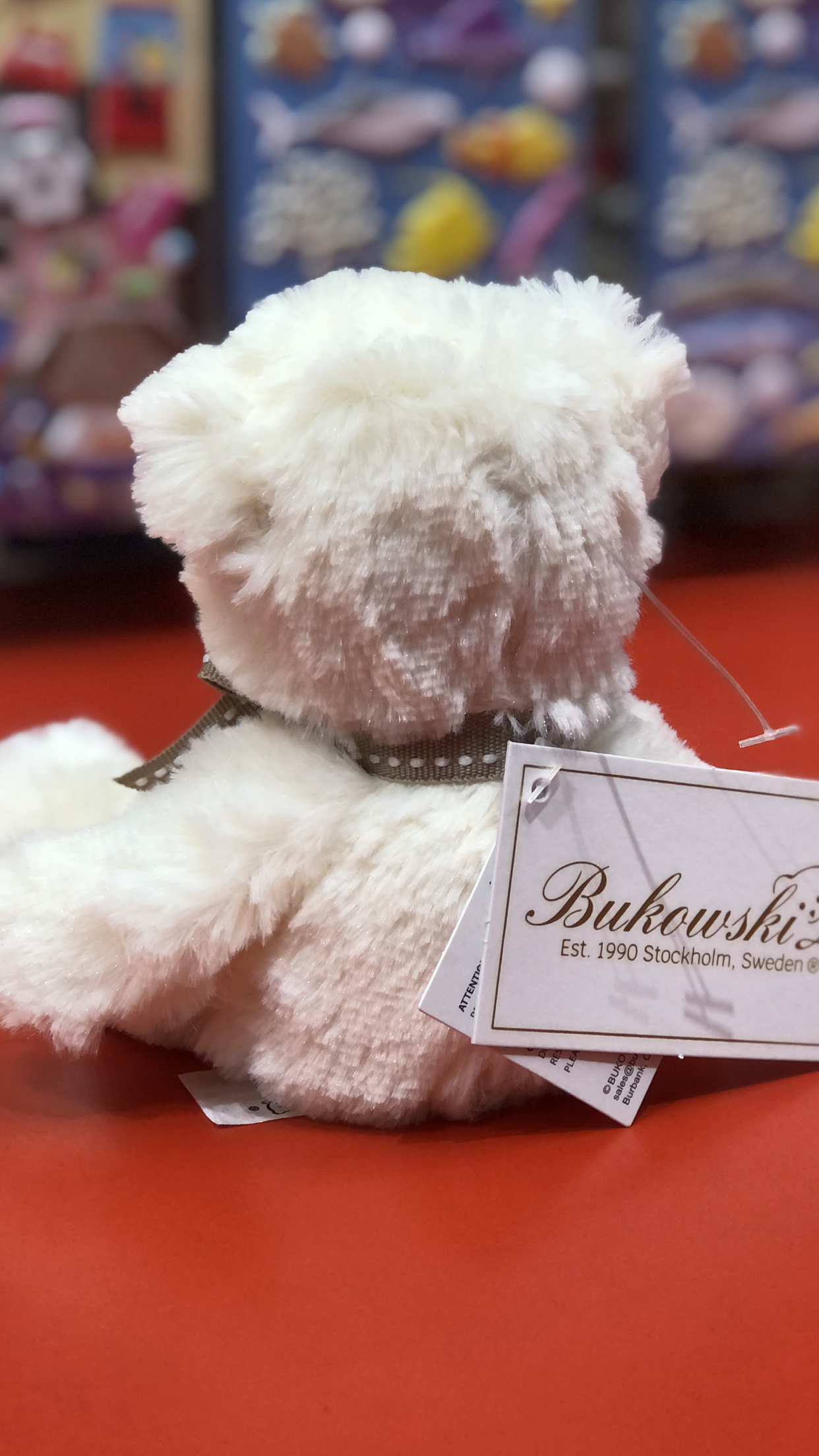 Soft white Bukowski bear plush toy Caspar with brand tag, sitting on red surface, colorful toy display background