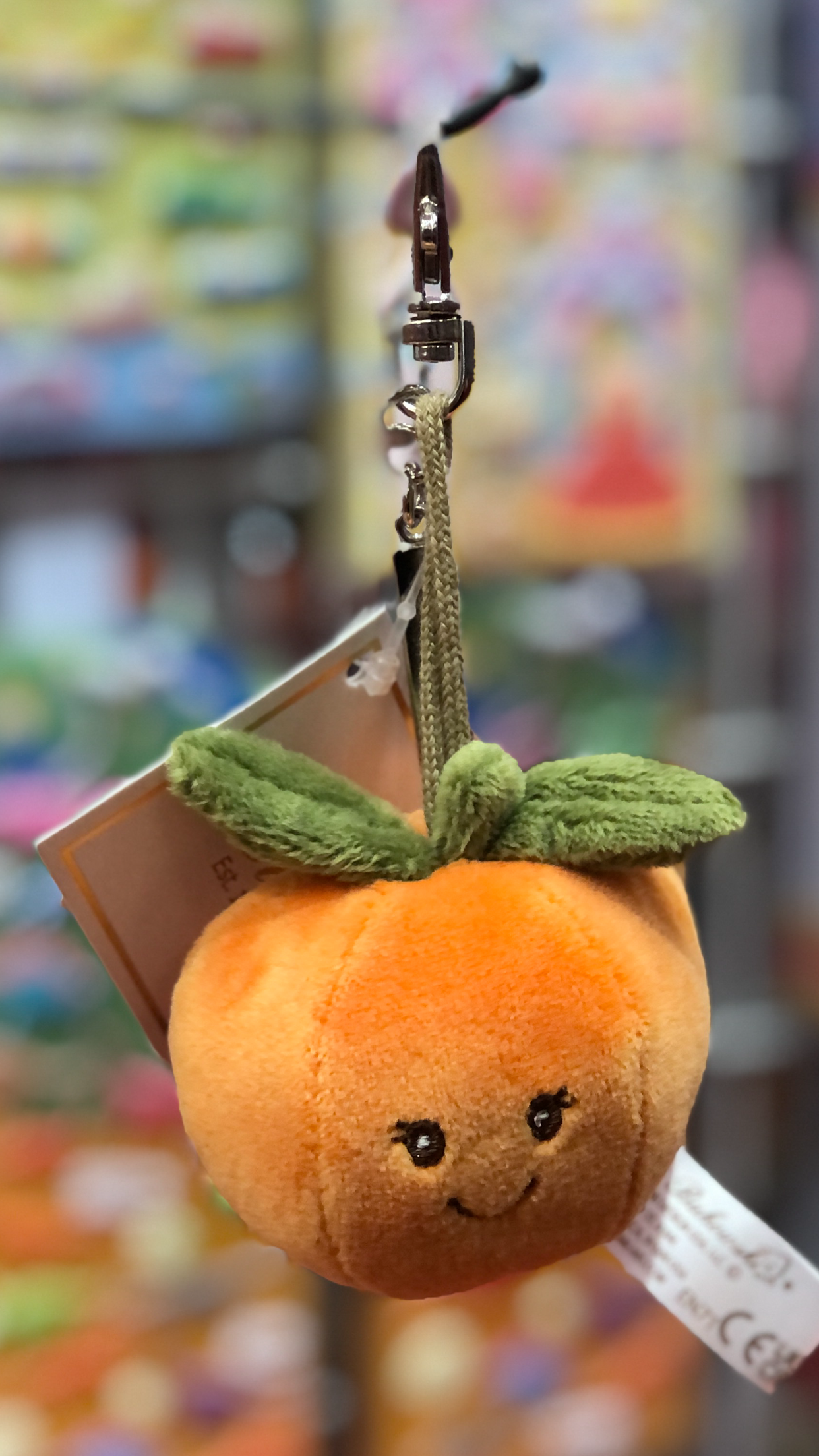 Cute plush orange keychain charm with smiling face and green leaf detail, hanging from clip against colorful background