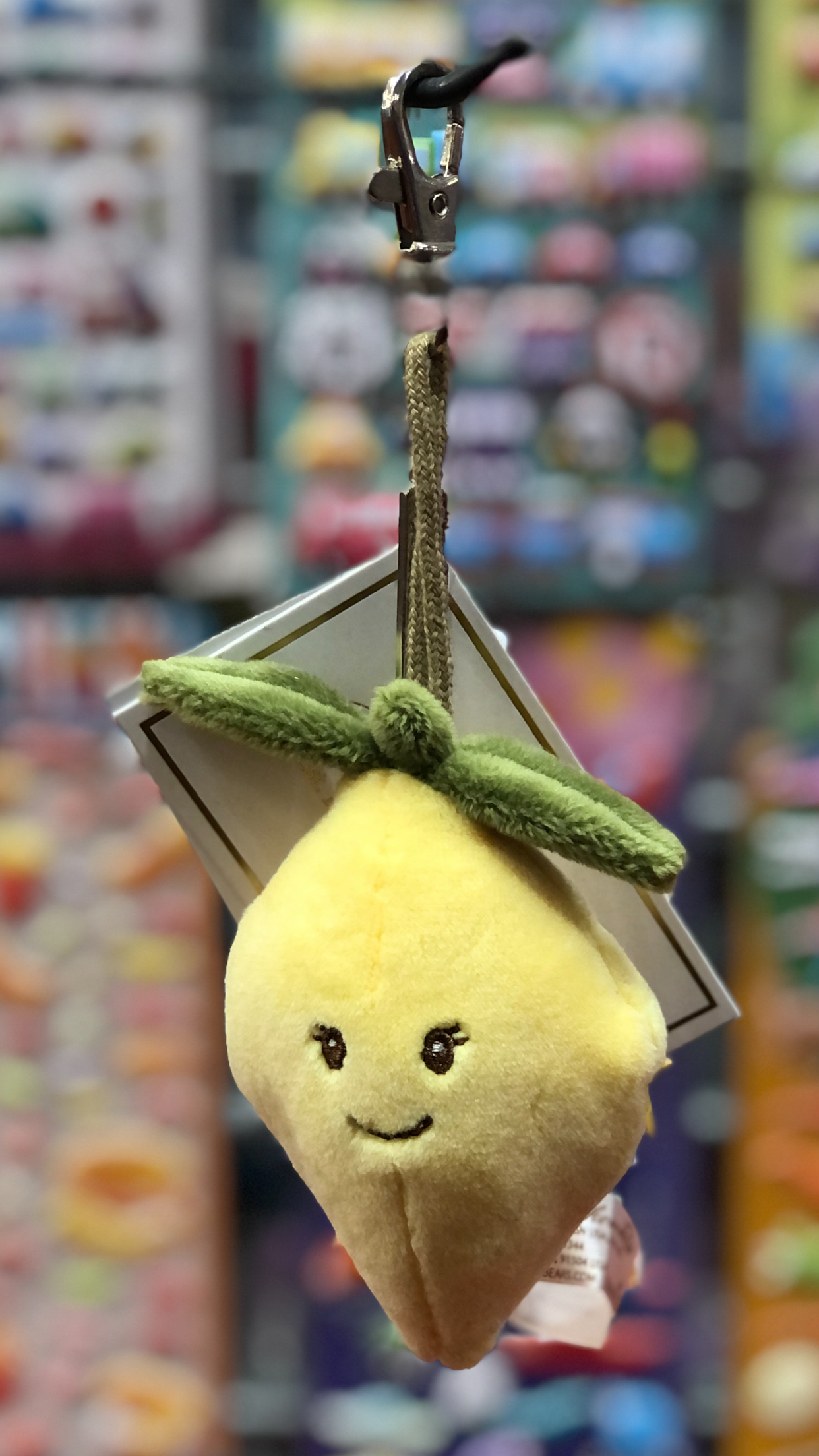Cute yellow lemon plush keyring with smiling face, hanging from carabiner clip against colorful blurred background