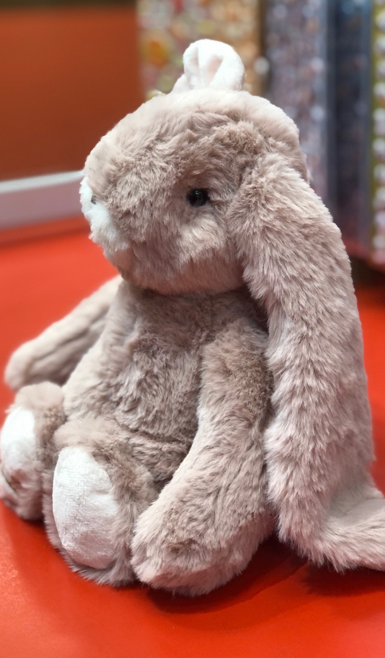 Soft pink Bukowski bunny plush toy with long ears and big eyes, sitting on red surface, comforting companion for children
