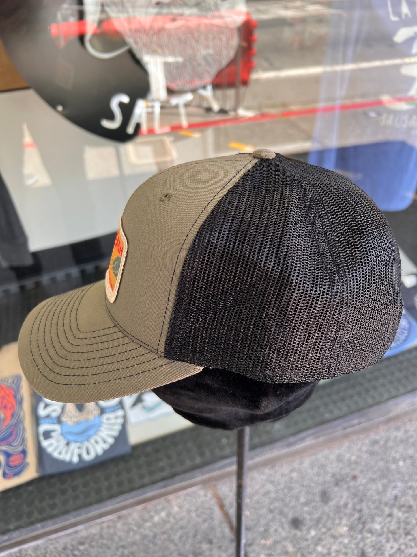 Olive Muir Beach trucker cap with mesh back, displaying patch logo, in store display window