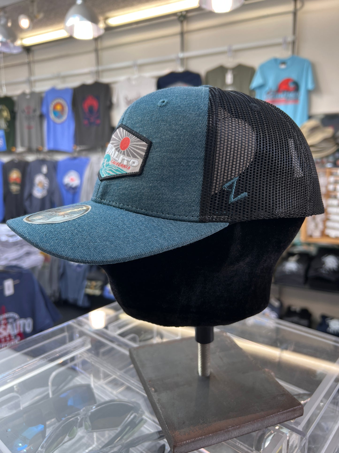Sausalito trucker cap with embroidered logo, teal front, black mesh back, displayed in souvenir shop with clothing racks
