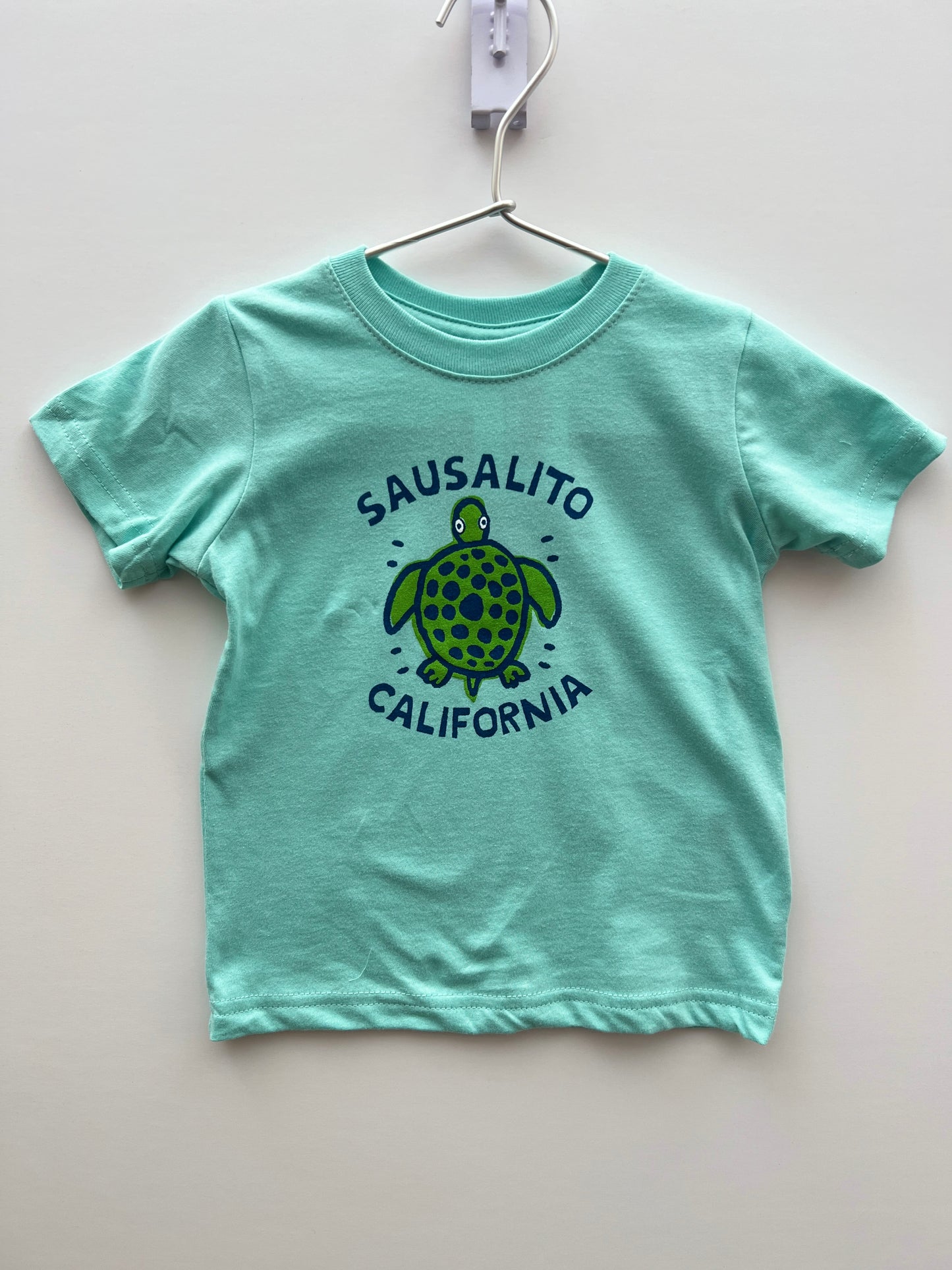 Mint green kids' t-shirt with cute turtle design and "Sausalito California" text, hanging on display