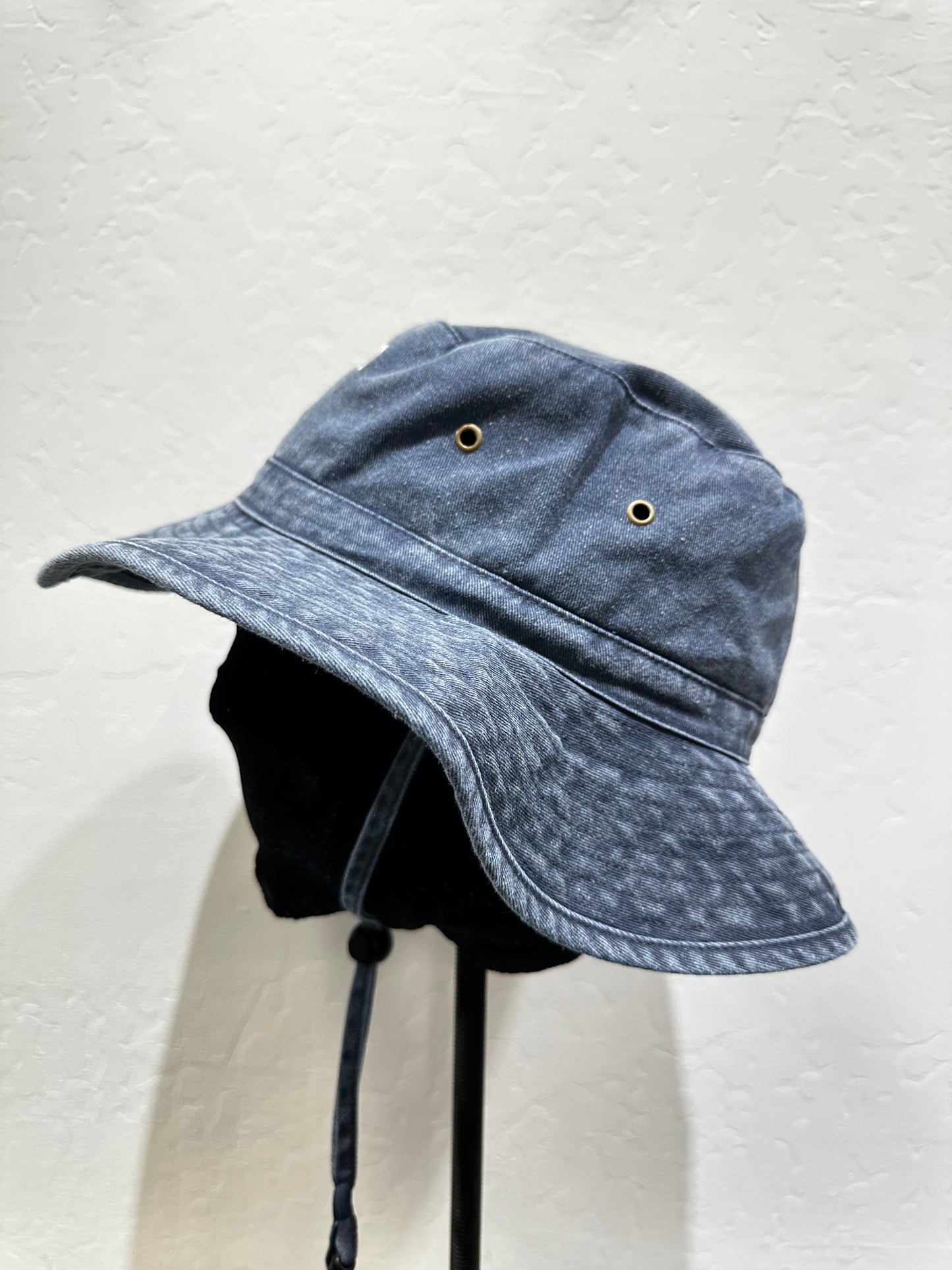 Washed cotton bucket hat in blue denim, wide brim, adjustable chin cord, displayed on stand against white background