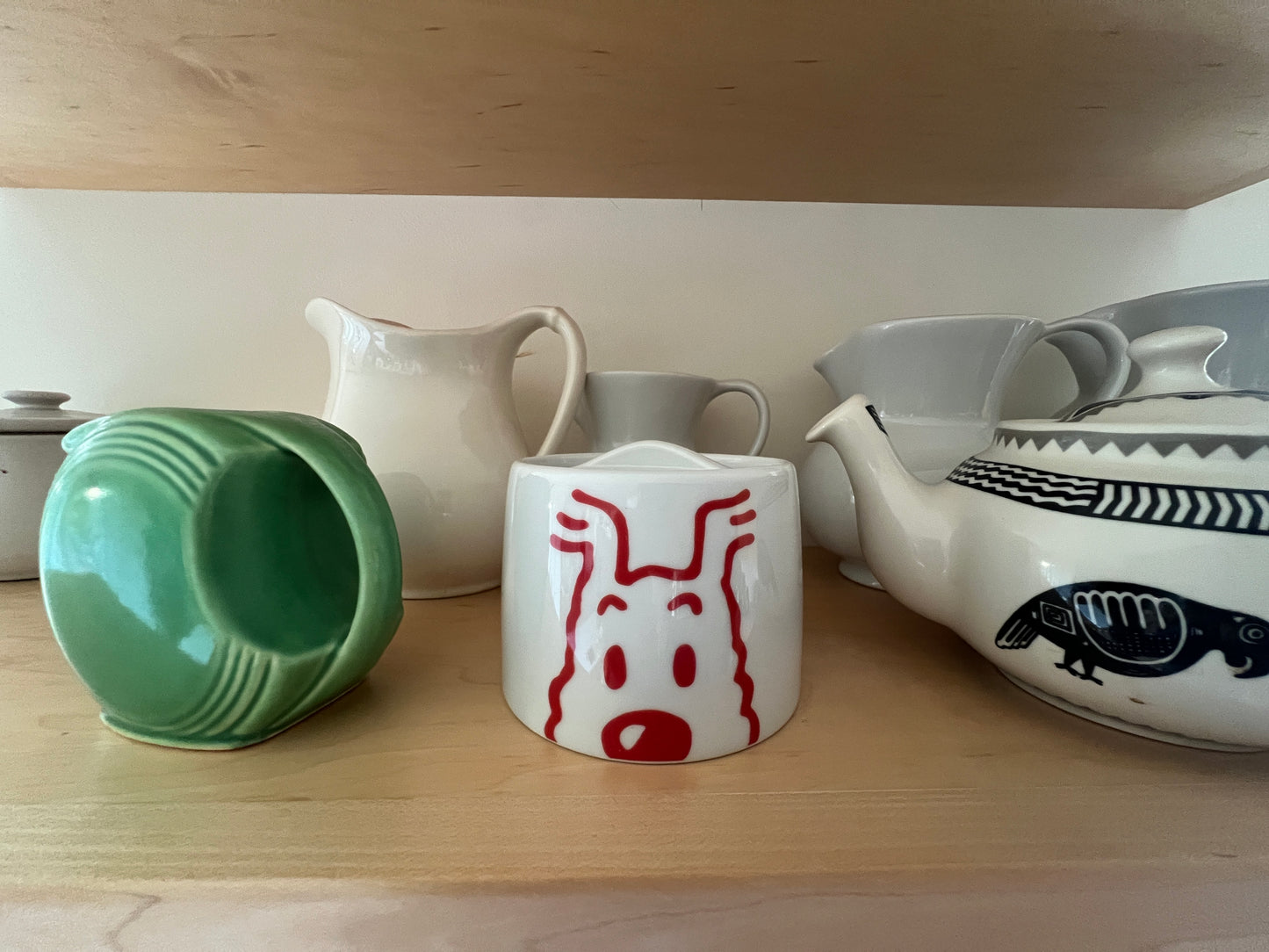 Whimsical porcelain sugar bowl with surprised face design, surrounded by other ceramic pieces on wooden shelf