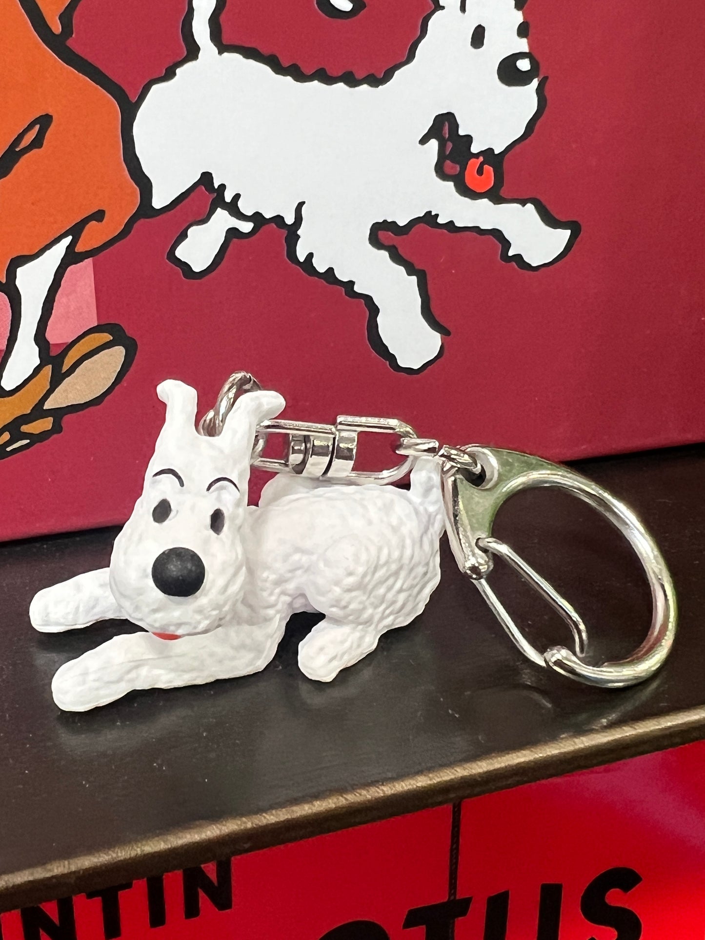 Snowy dog keychain figure, 4cm long, laying down with metal ring, displayed with cartoon backdrop