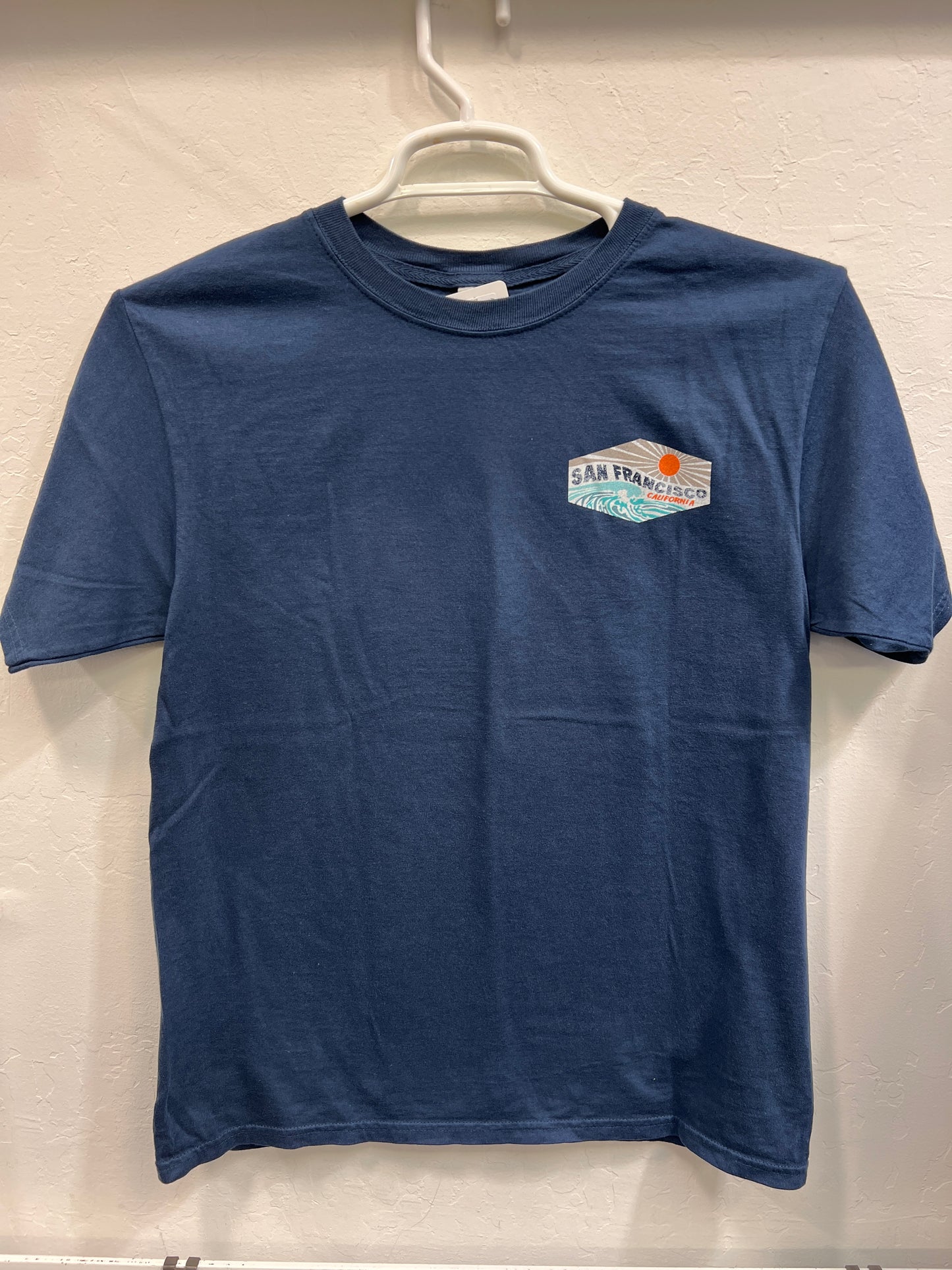 Navy Affirmation Wave unisex t-shirt with San Francisco wave logo, hanging on white hanger against beige wall
