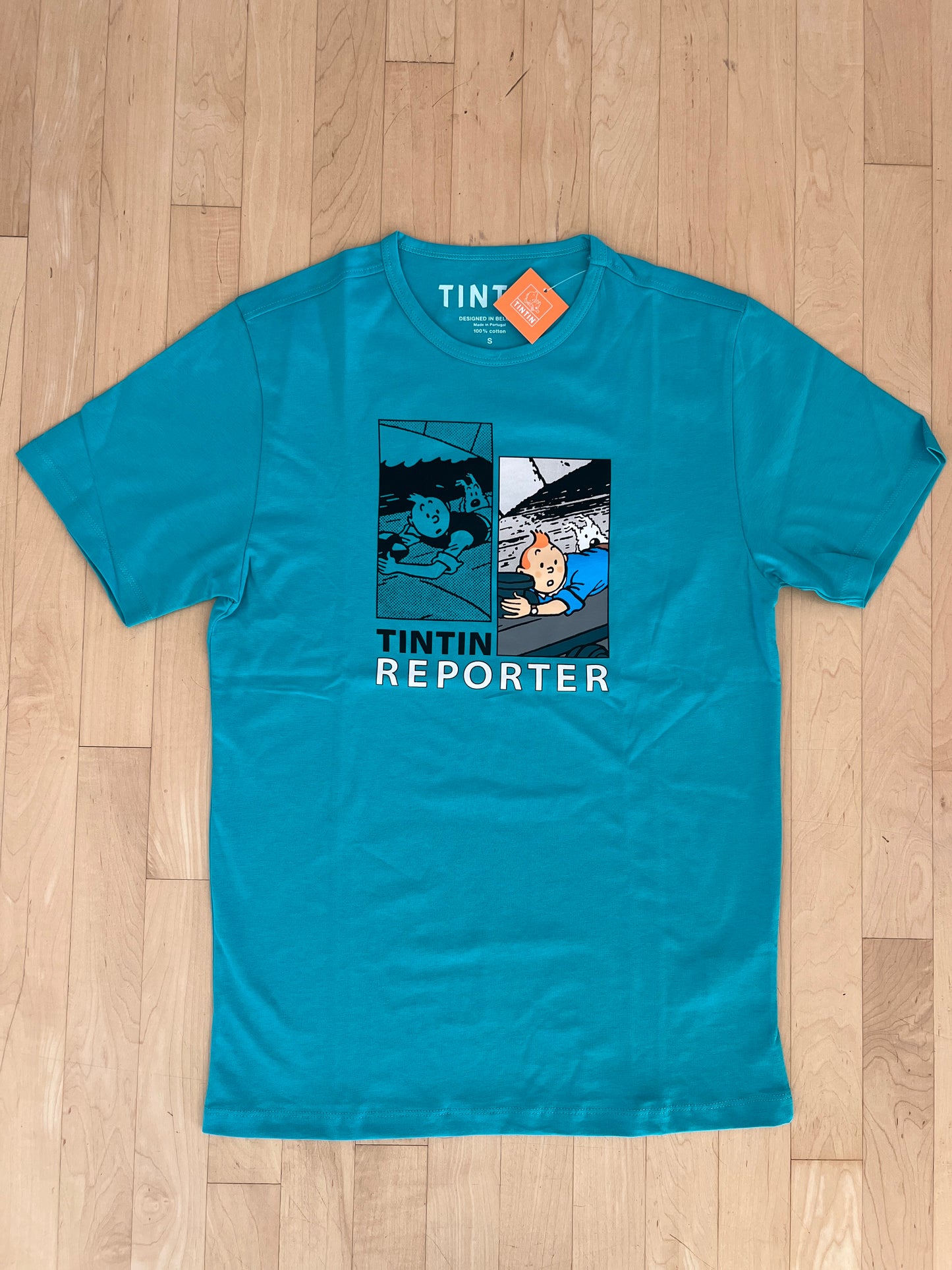 Turquoise Tintin Reporter t-shirt with comic panels showing character on train roof, laid on wooden floor