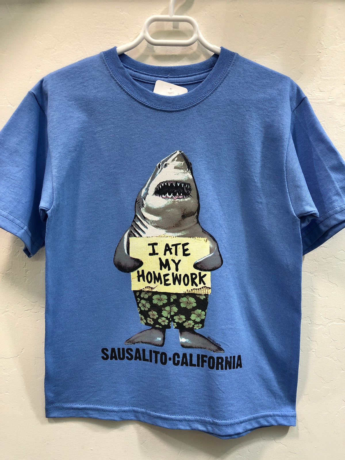 Blue kids' t-shirt with cartoon shark holding "I ate my homework" sign, Sausalito California printed below