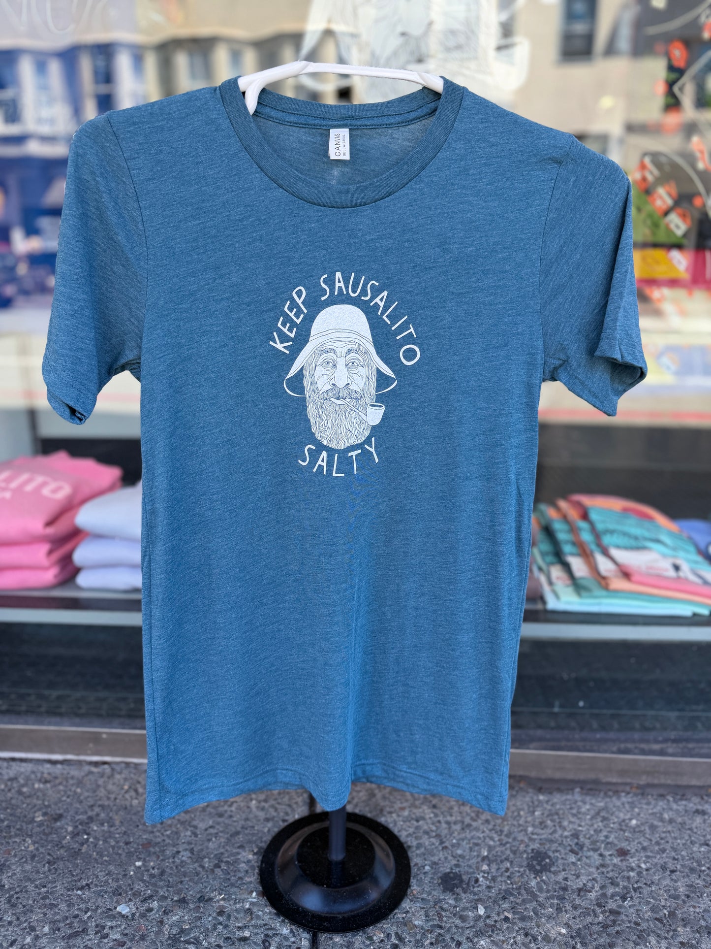 Front view of a steel blue, heathered crewneck t-shirt displayed on a black tabletop clothing mannequin stand in front of a shop window. The shirt features a white circular graphic screen-printed on the center chest, showcasing a detailed illustration of a classic, bearded coastal fisherman wearing a traditional weather hat and smoking a pipe. The text "KEEP SAUSALITO" arches cleanly over the top of the portrait, and the word "SALTY" curves directly underneath.