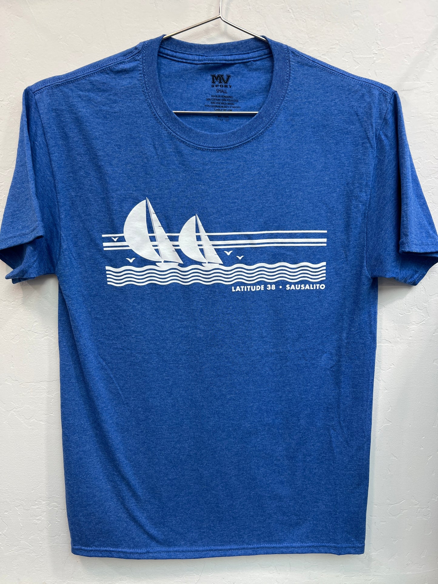 A heather blue crewneck t-shirt hanging on a wire hanger against a white textured wall. The shirt features a white screen-printed graphic across the chest depicting two sailboats with full spinnakers sailing on stylized ocean waves under a three-line horizon, accompanied by the text "LATITUDE 38 • SAUSALITO" in a clean sans-serif font.