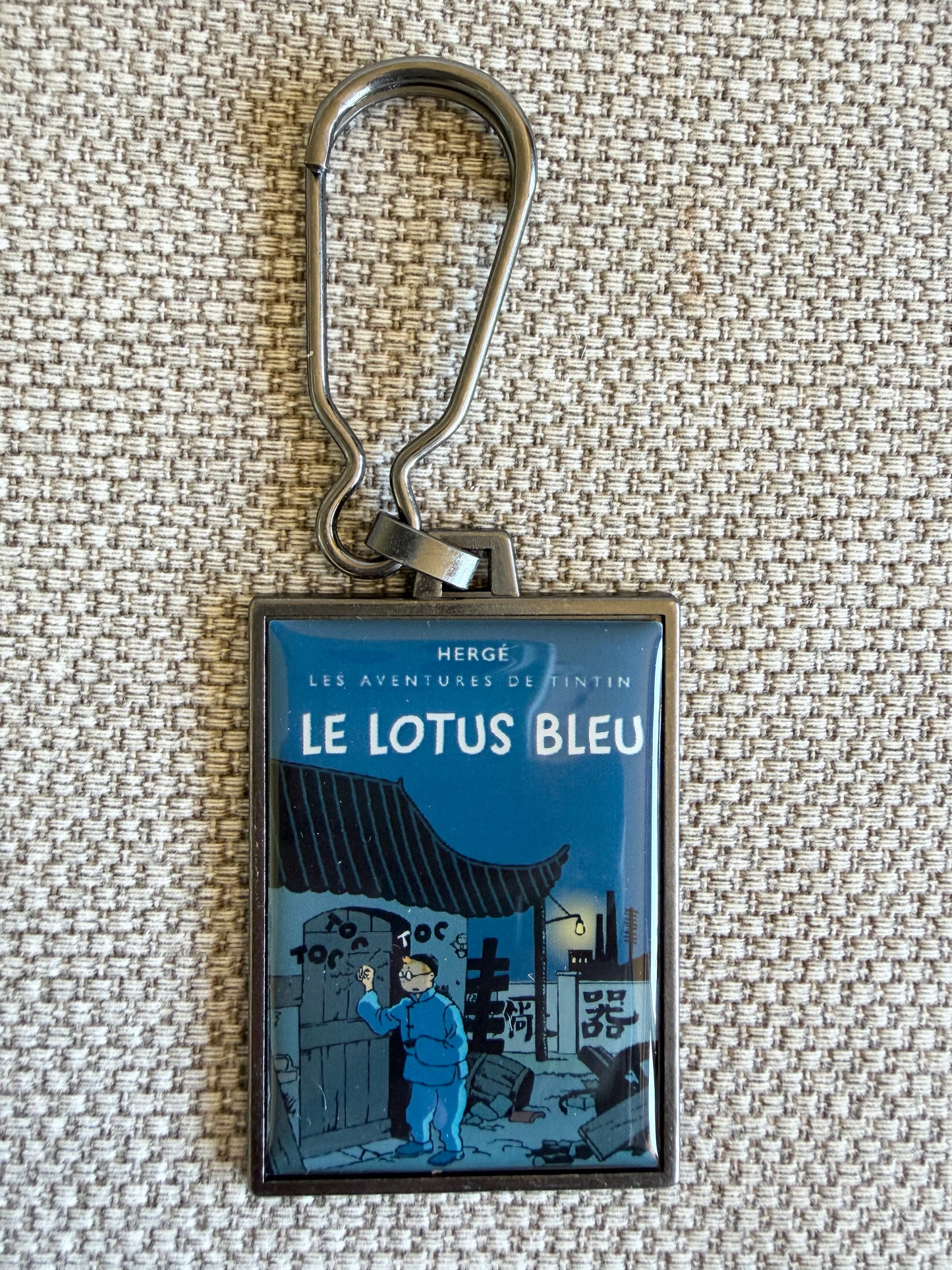 A rectangular keychain displaying the newly colorized book cover art for Hergé's Tintin adventure "Le Lotus Bleu." The atmospheric illustration shows Tintin wearing blue Chinese robes, hiding inside a large porcelain vase alongside Snowy, while a stylized black dragon looms on a vibrant red wall behind them in a dark Shanghai night scene.
