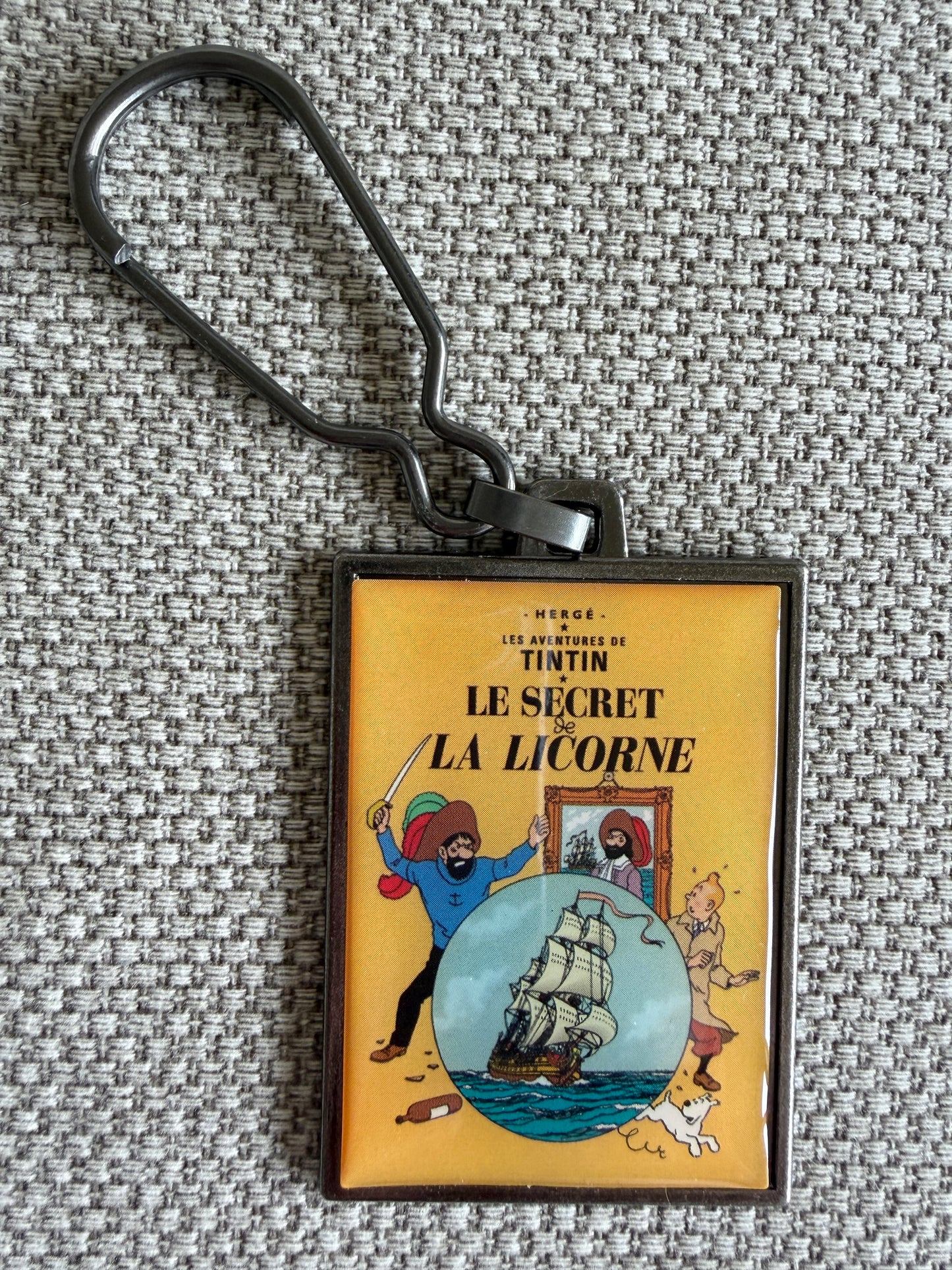 A close-up photograph of a rectangular metal keychain resting on a textured, grey woven fabric background. The keychain displays the classic French cover art for Hergé's Tintin adventure "Le Secret de La Licorne," showing Tintin standing behind an image of the 17th-century sailing ship, The Unicorn, against a warm golden-yellow backdrop.