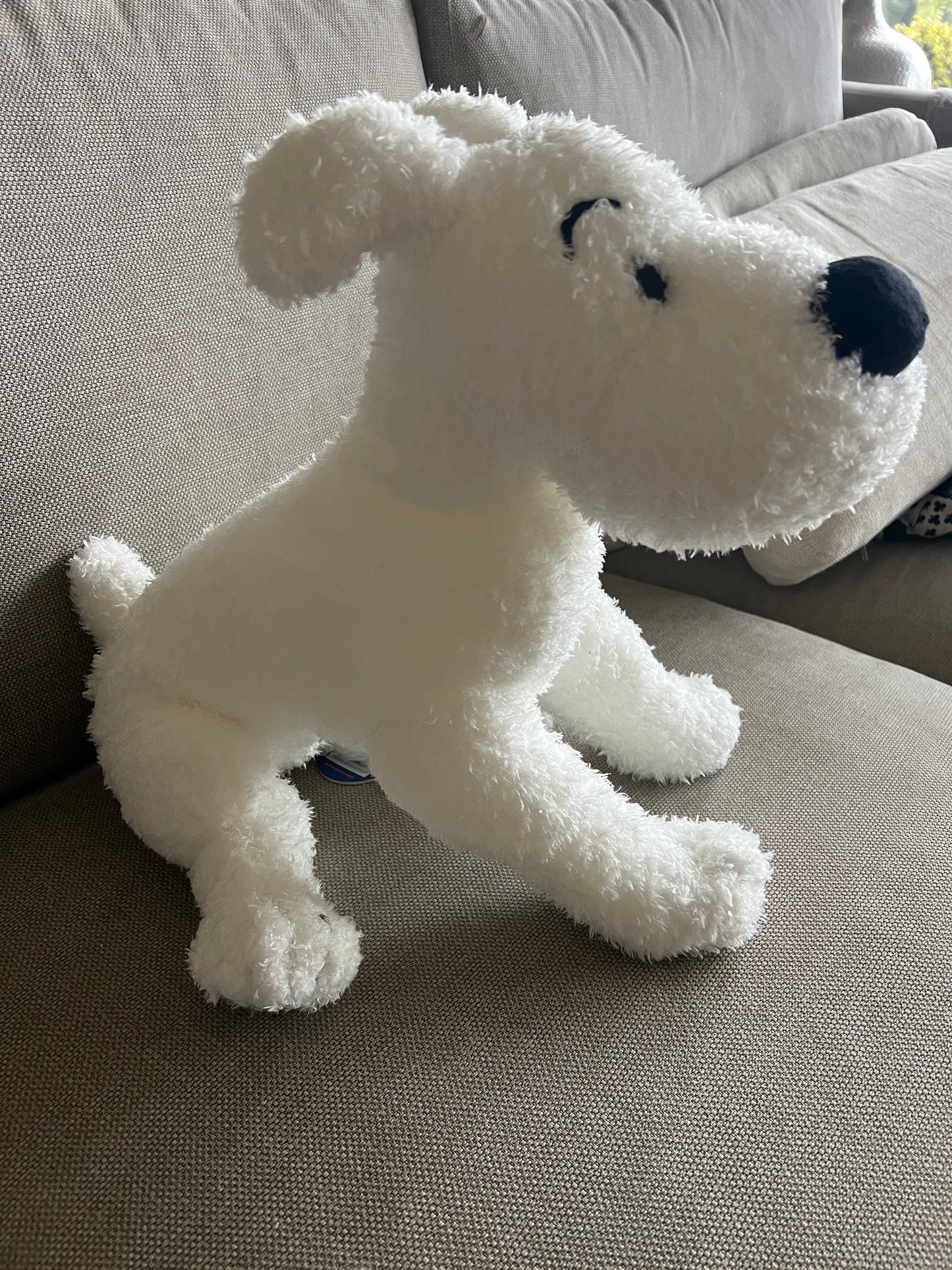 A side profile view of a large, fluffy white plush Snowy dog sitting upright on a textured grey fabric couch. The plush shows a soft, crimped faux-fur texture, a small pointed tail, and distinct black embroidered detail for its closed eye and arched eyebrow.