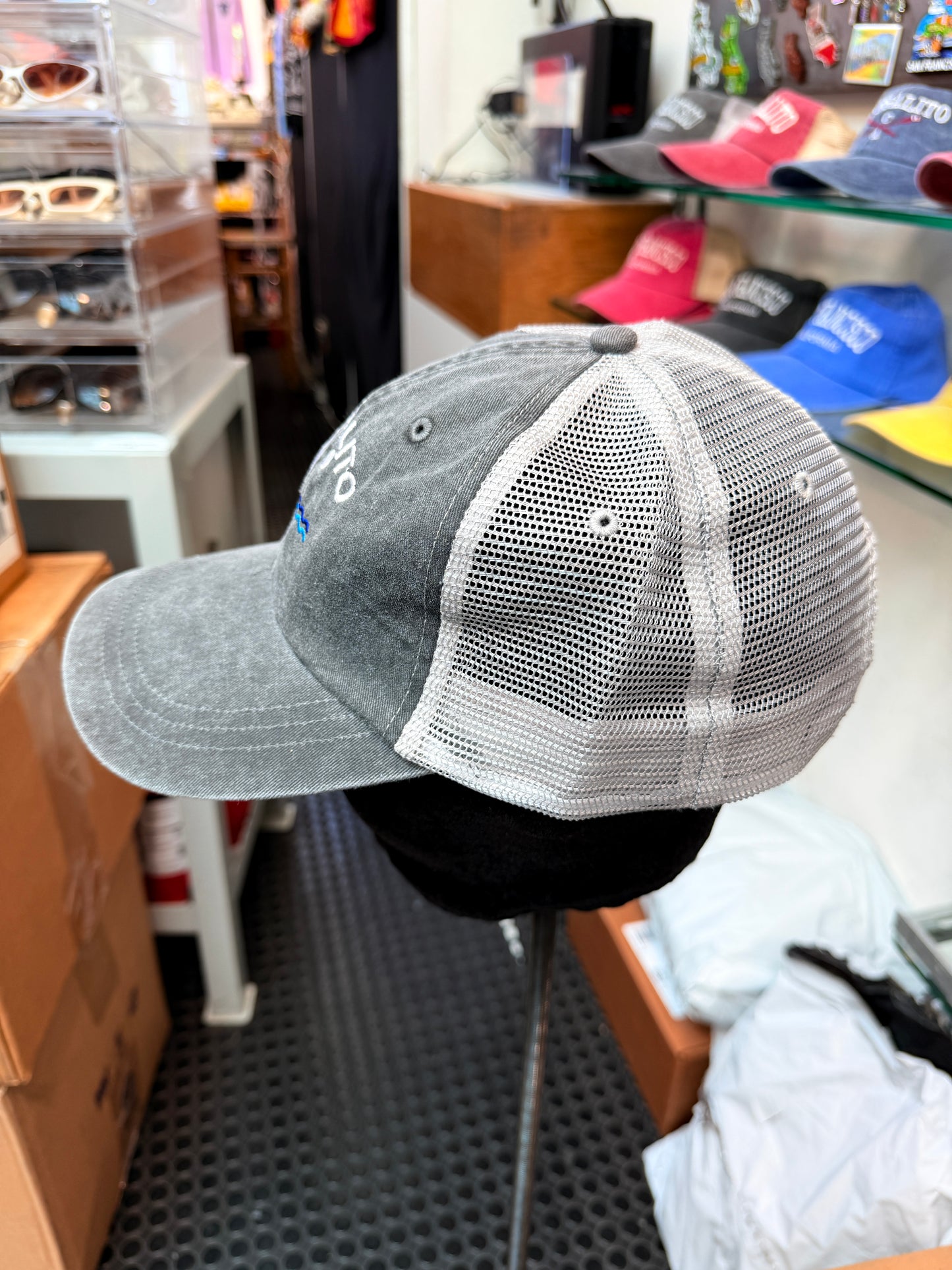 A side profile view of an unstructured trucker hat on a display stand inside a shop. The front panels and brim are made of a washed, pigment-dyed dark grey cotton twill, while the back sections feature a matching grey breathable mesh. Crisp white embroidered block lettering that reads "SAUSALITO" curves across the front over a detailed pair of crossed rowing sculls.